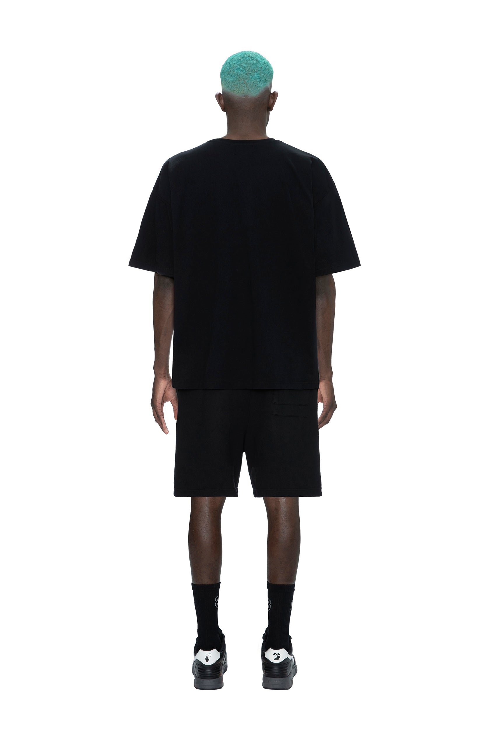 A person with teal hair, dressed in the 6PM PAID COLLAB T-SHIRT BLACK, black shorts, socks, and sneakers, stands facing away on a white background.