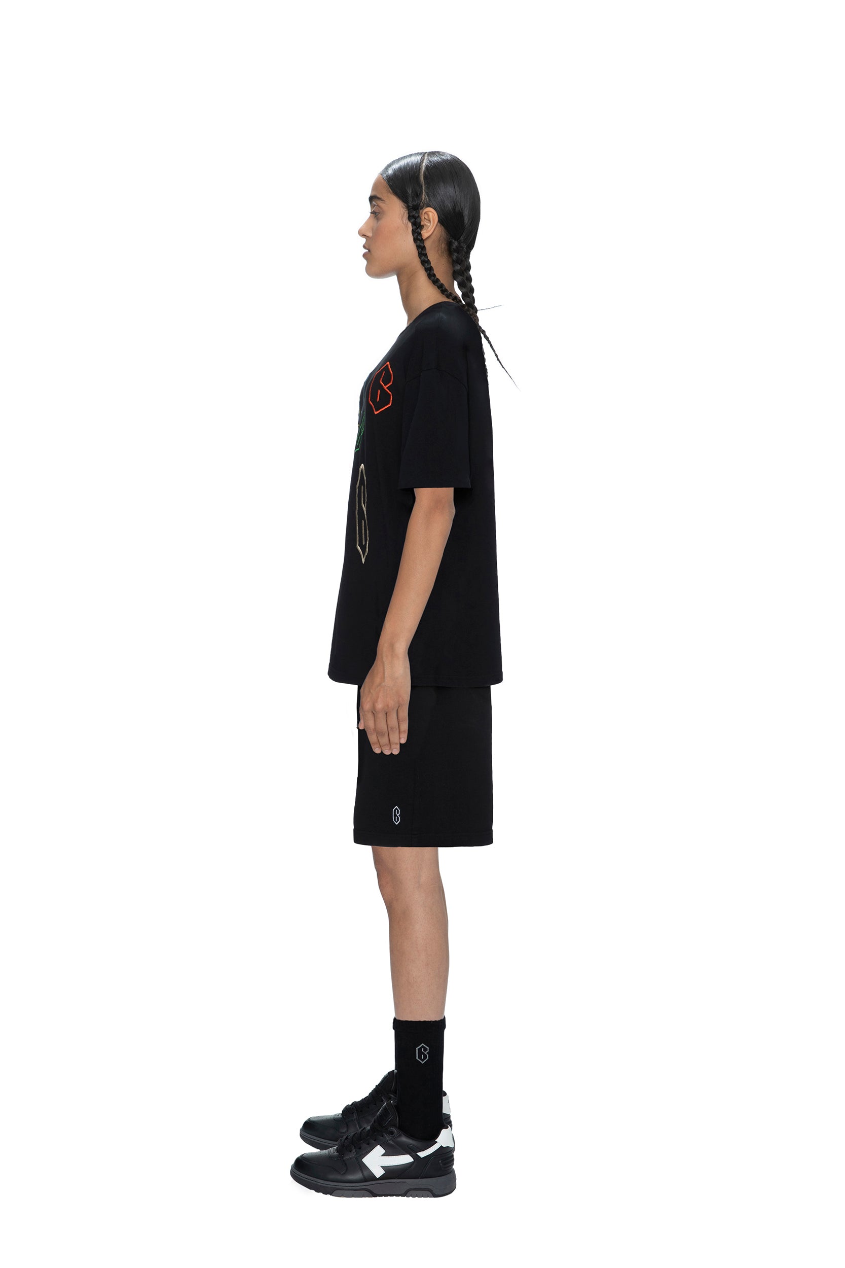 A person with long braided hair stands in profile against a white background, wearing the 6PM SUPER 6 T-SHIRT in black, paired with black shorts, socks, and sneakers.