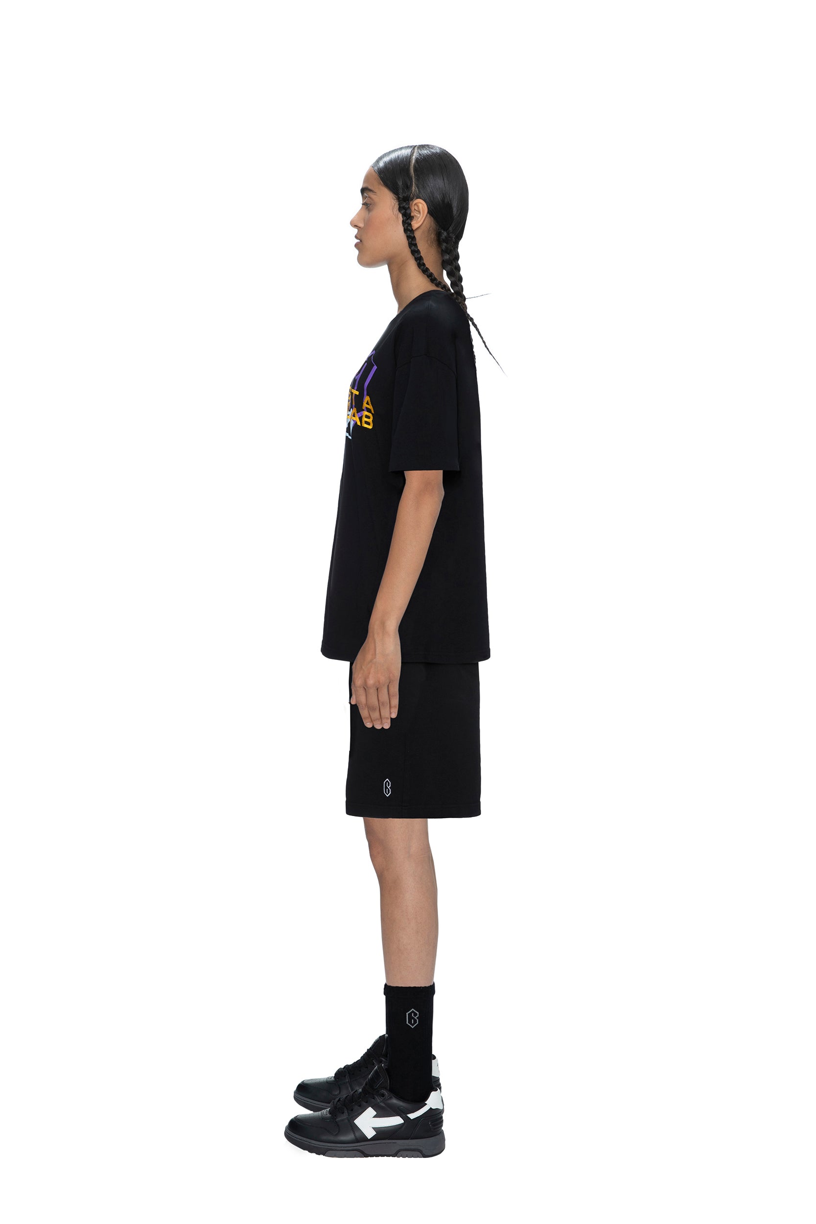 A person with long, dark braids stands in profile against a white background, wearing the 6PM PAID COLLAB T-SHIRT BLACK, along with black shorts, black crew socks, and black sneakers.