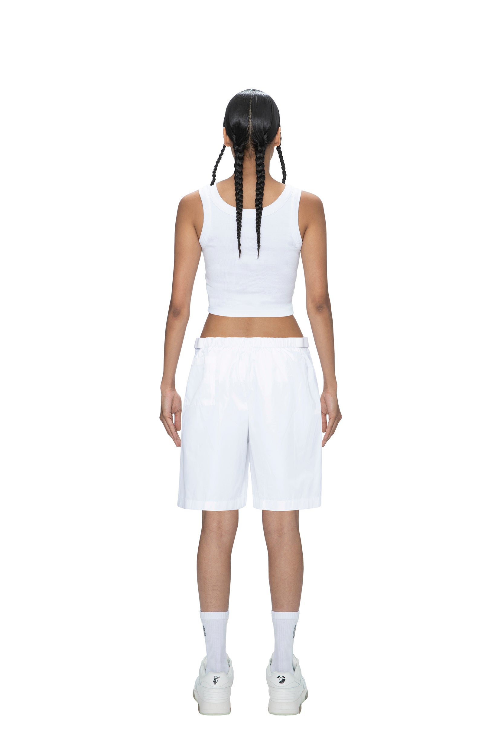A person with two long braids, wearing 6PM TECH SHORTS WHITE, a white tank top, white socks, and white sneakers, stands facing away on a plain white background.