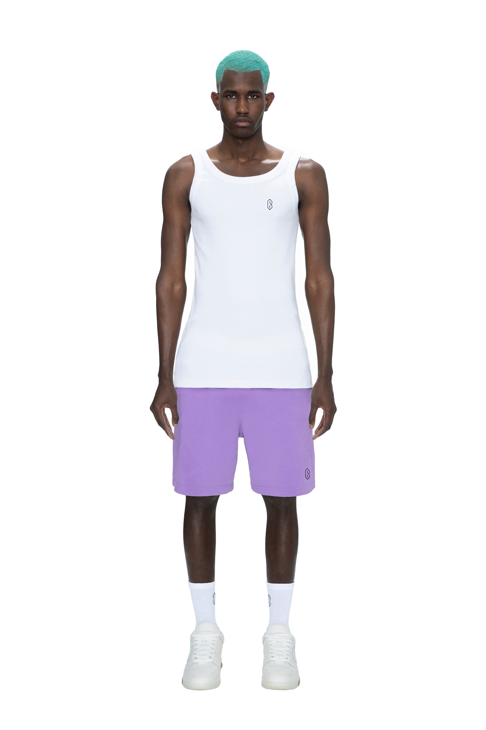 A man with teal hair wears a white tank top, 6PM SWEATSHORTS LILAC, white socks, and white sneakers, standing facing forward against a plain white background.