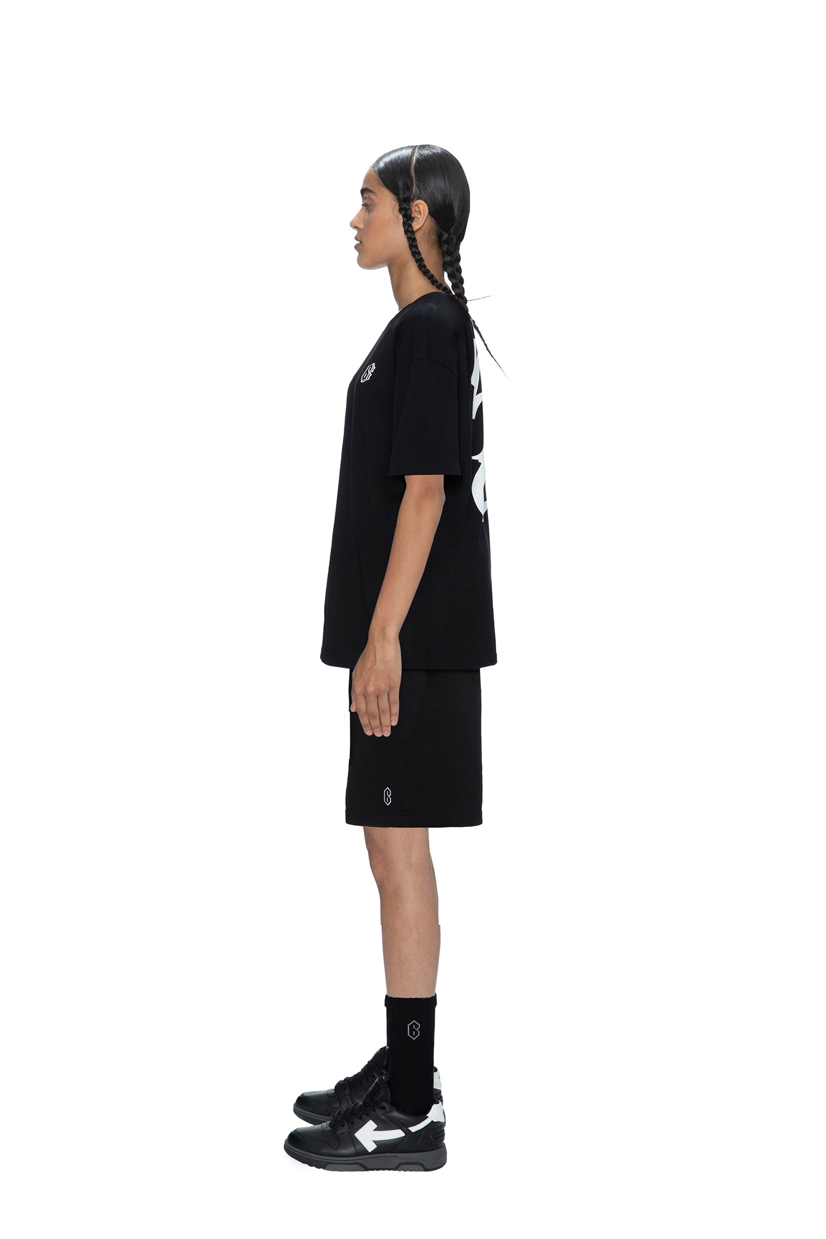 A person with long hair in two braids stands in profile against a white background, wearing the 6PM SIGNATURE T-SHIRT BLACK, paired with black shorts, socks, and sneakers.