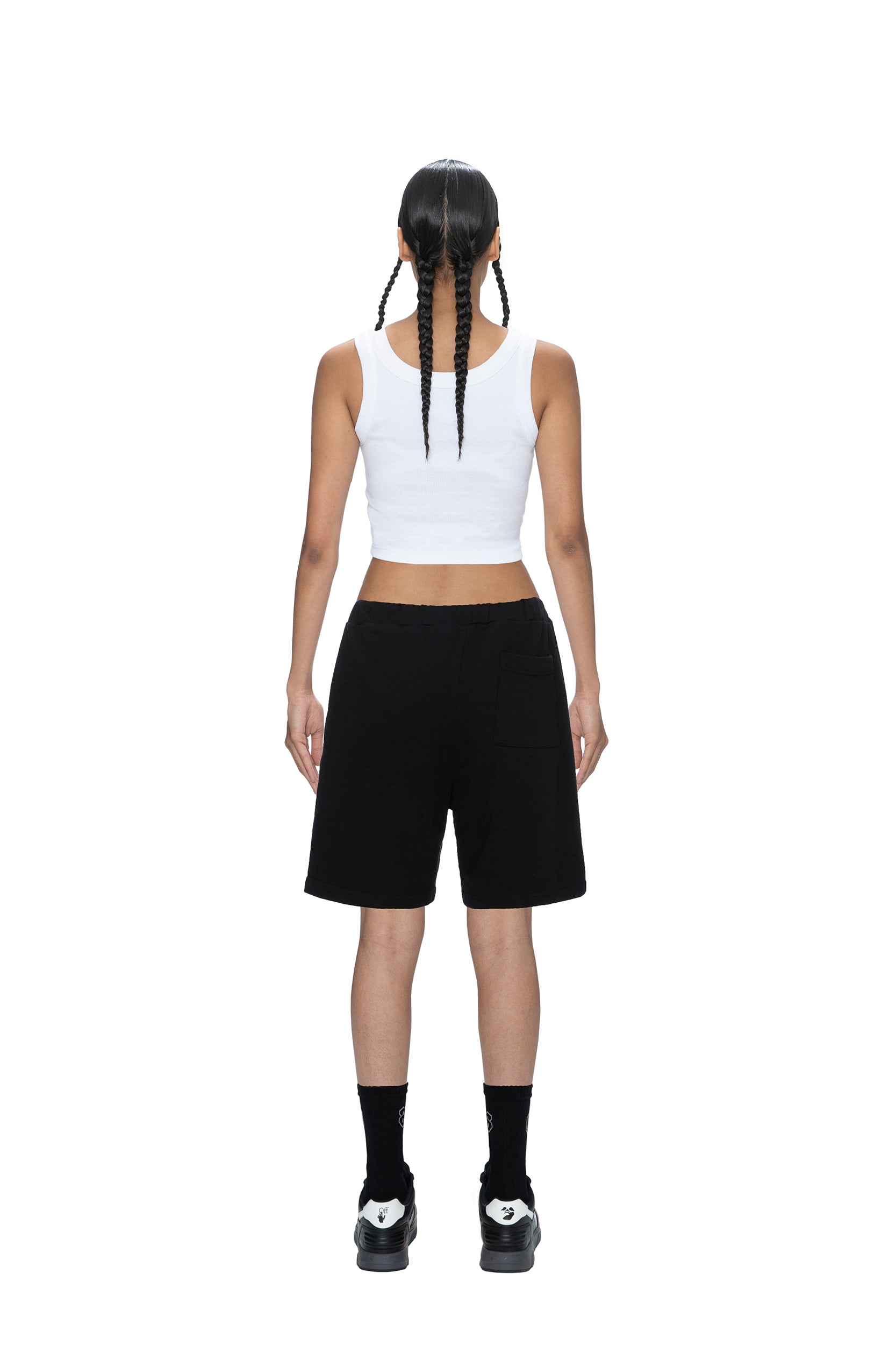 A person with two long braids stands facing away, wearing a white sleeveless crop top, 6PM SWEATSHORTS BLACK, black socks, and black shoes against a plain white background.