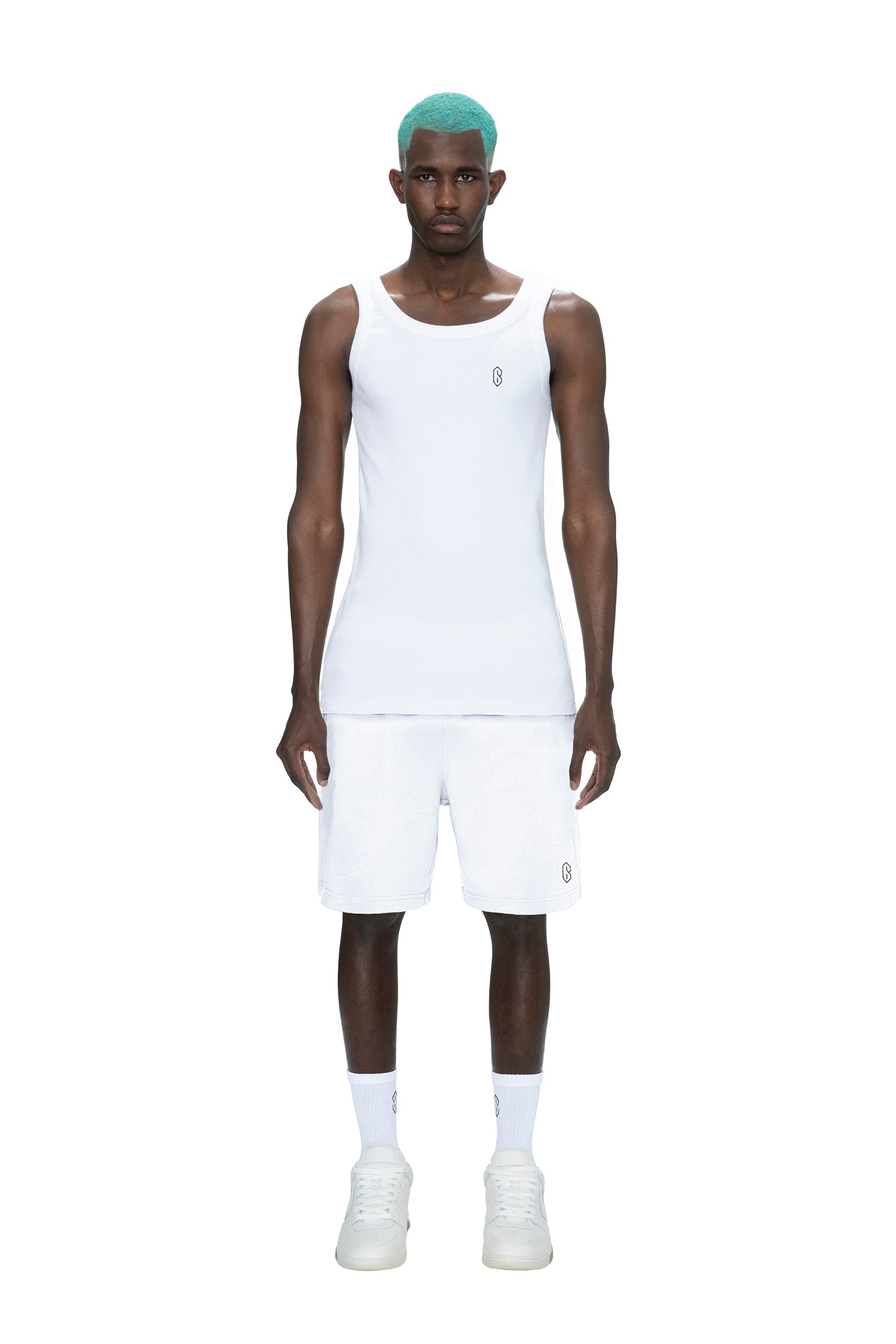A man with turquoise hair wears 6PM SWEATSHORTS WHITE and a white tank top, socks, and sneakers, standing against a plain white background.