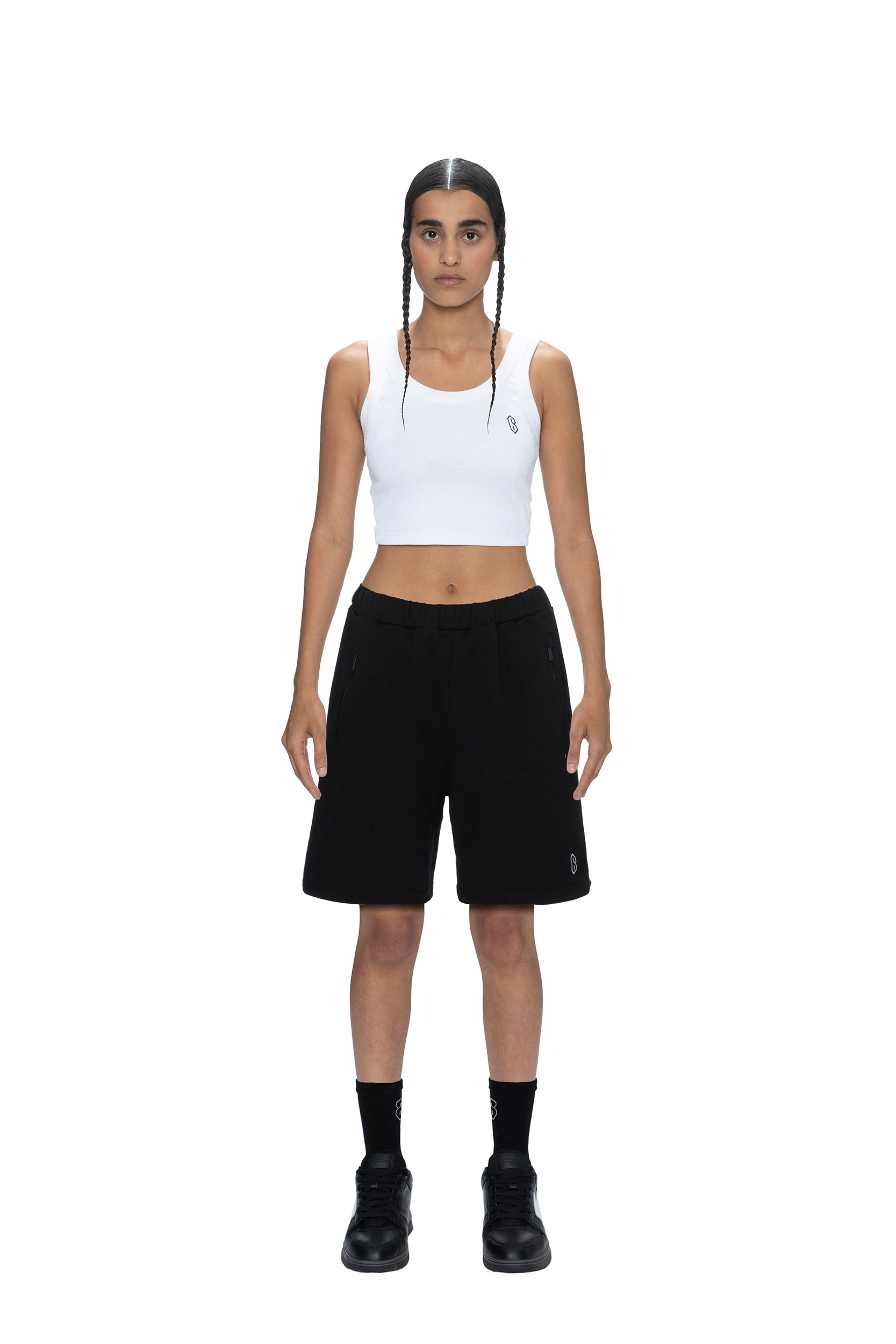 A person with long, dark braided hair wears 6PM SWEATSHORTS BLACK, a white crop tank top, black socks, and black shoes, standing facing forward against a plain white background.