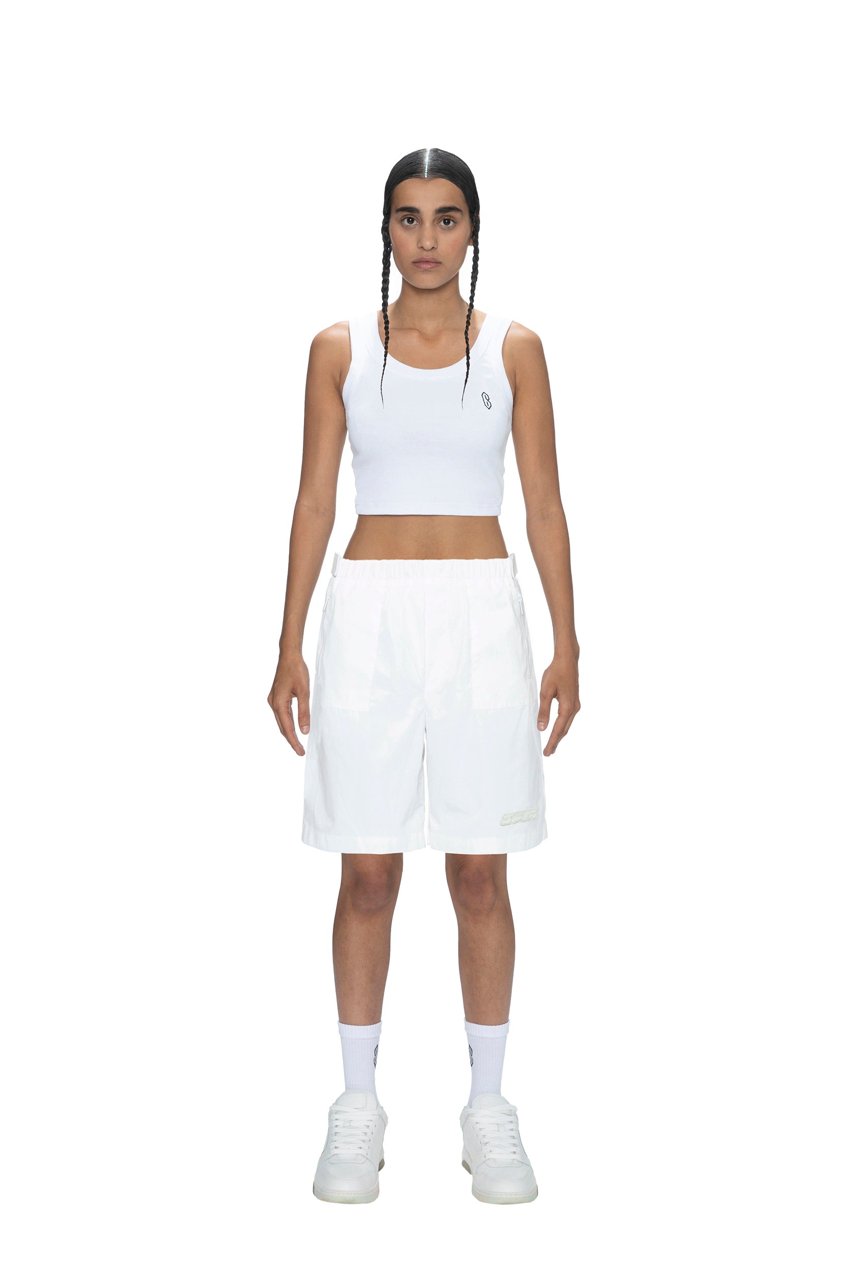 A person with braided hair wears 6PM’s TECH SHORTS WHITE, a white crop top, white socks, and white sneakers, standing against a plain white background, facing forward with arms relaxed at their sides.
