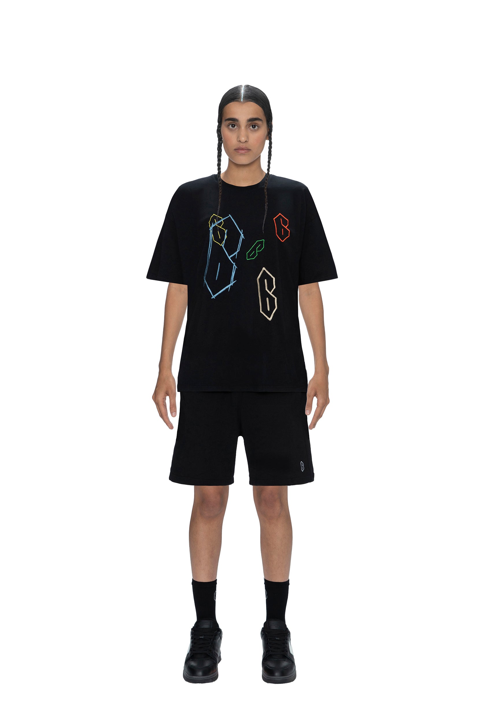 A person with two braids stands facing forward, wearing the 6PM SUPER 6 T-SHIRT BLACK with colorful abstract shapes, paired with black shorts, socks, and shoes against a plain white background.