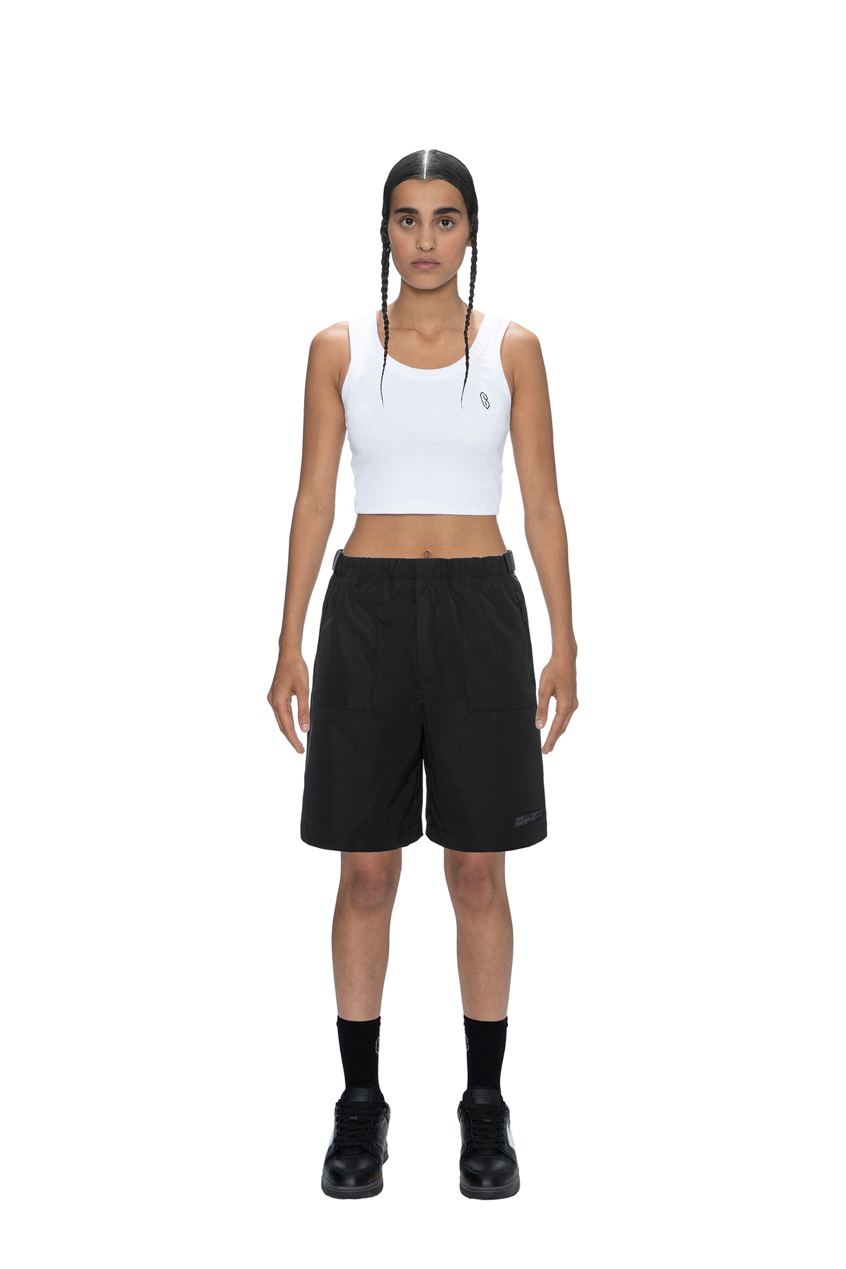 A person with braided hair wears black 6PM TECH SHORTS, a white cropped tank top, black socks, and black shoes, standing against a plain white background.