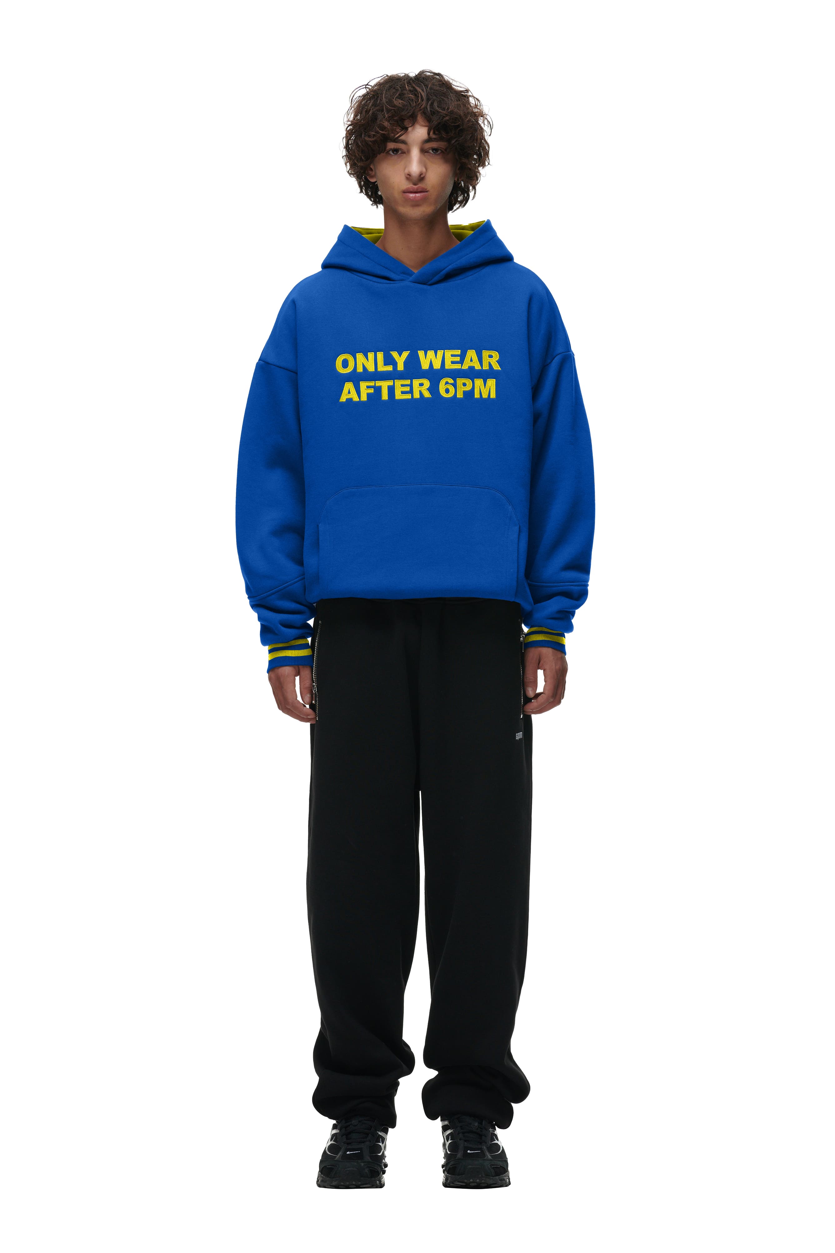 A person with curly hair wears the 6PM ONLY WEAR HOODIE BLUE with yellow lettering, styled with black pants and shoes, and stands against a plain white background.