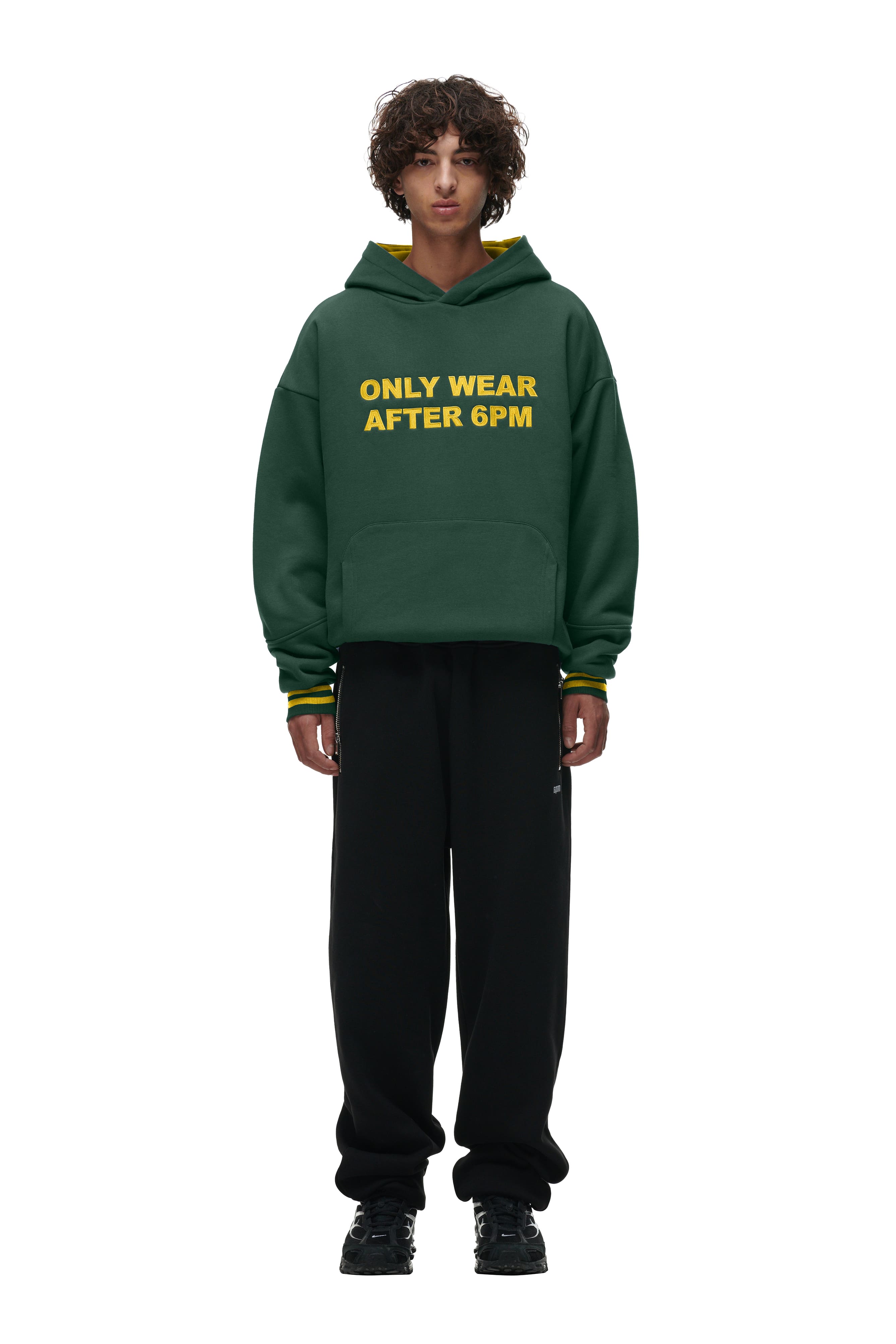 A person faces forward against a white background, wearing the 6PM ONLY WEAR HOODIE GREEN with bold yellow text, paired with black pants and black shoes.