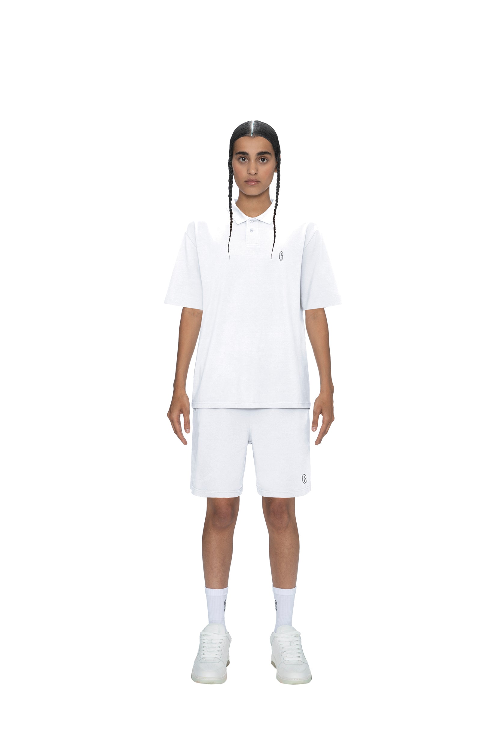 A person with long braids stands facing forward, wearing the 6PM POLO SHIRT WHITE, white shorts, and white sneakers against a plain white background.