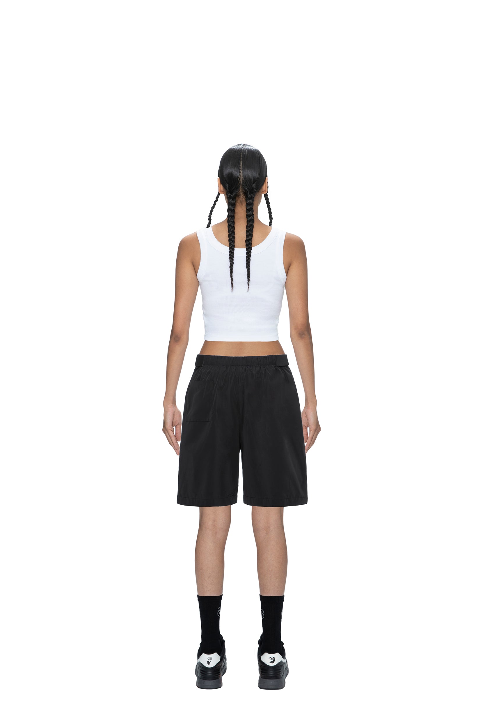 A person with two long braids stands with their back to the camera, wearing a white tank top, 6PM TECH SHORTS BLACK, black socks, and black-and-white sneakers against a plain white background.