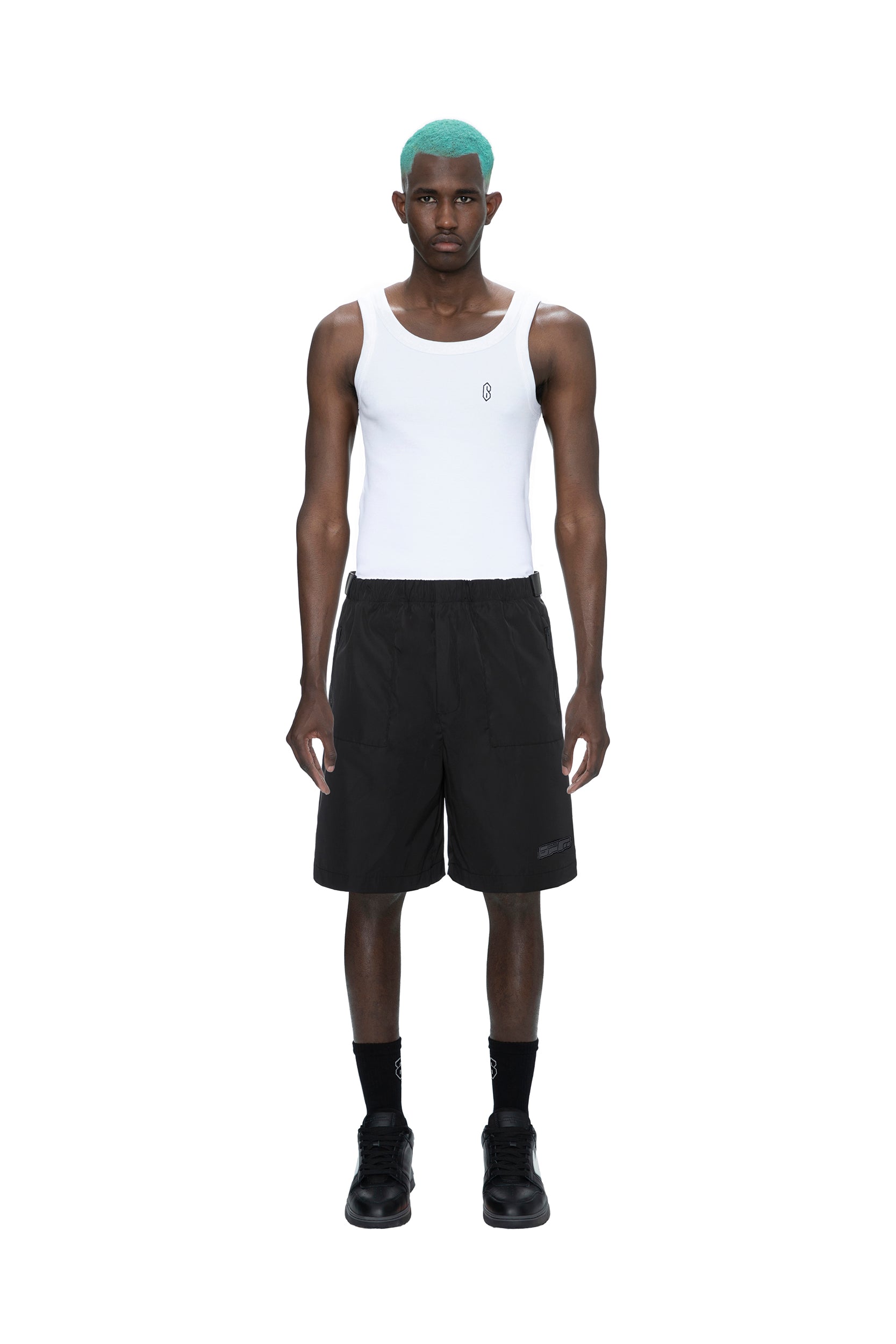 A man with teal hair wears a white tank top, 6PM TECH SHORTS BLACK, black socks, and black shoes, standing against a plain white background.