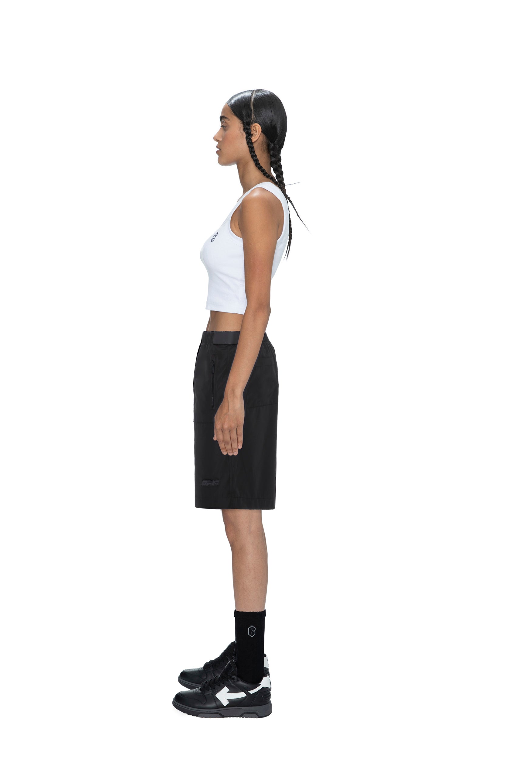 A woman with braided hair stands in profile against a white background, wearing a white crop top, black socks, black sneakers, and 6PM TECH SHORTS BLACK.