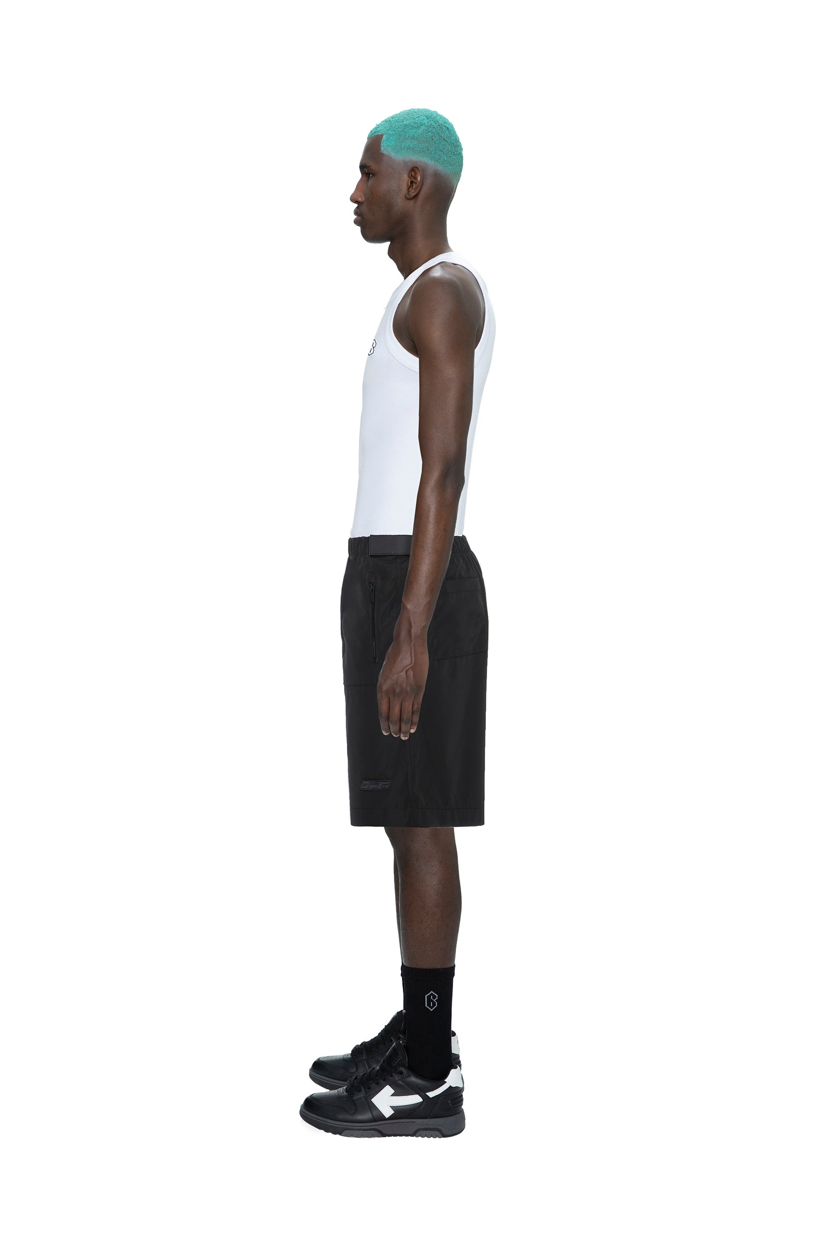 A man with turquoise hair, dressed in a white tank top, 6PM TECH SHORTS BLACK, black socks, and black sneakers, stands in profile against a plain white background.