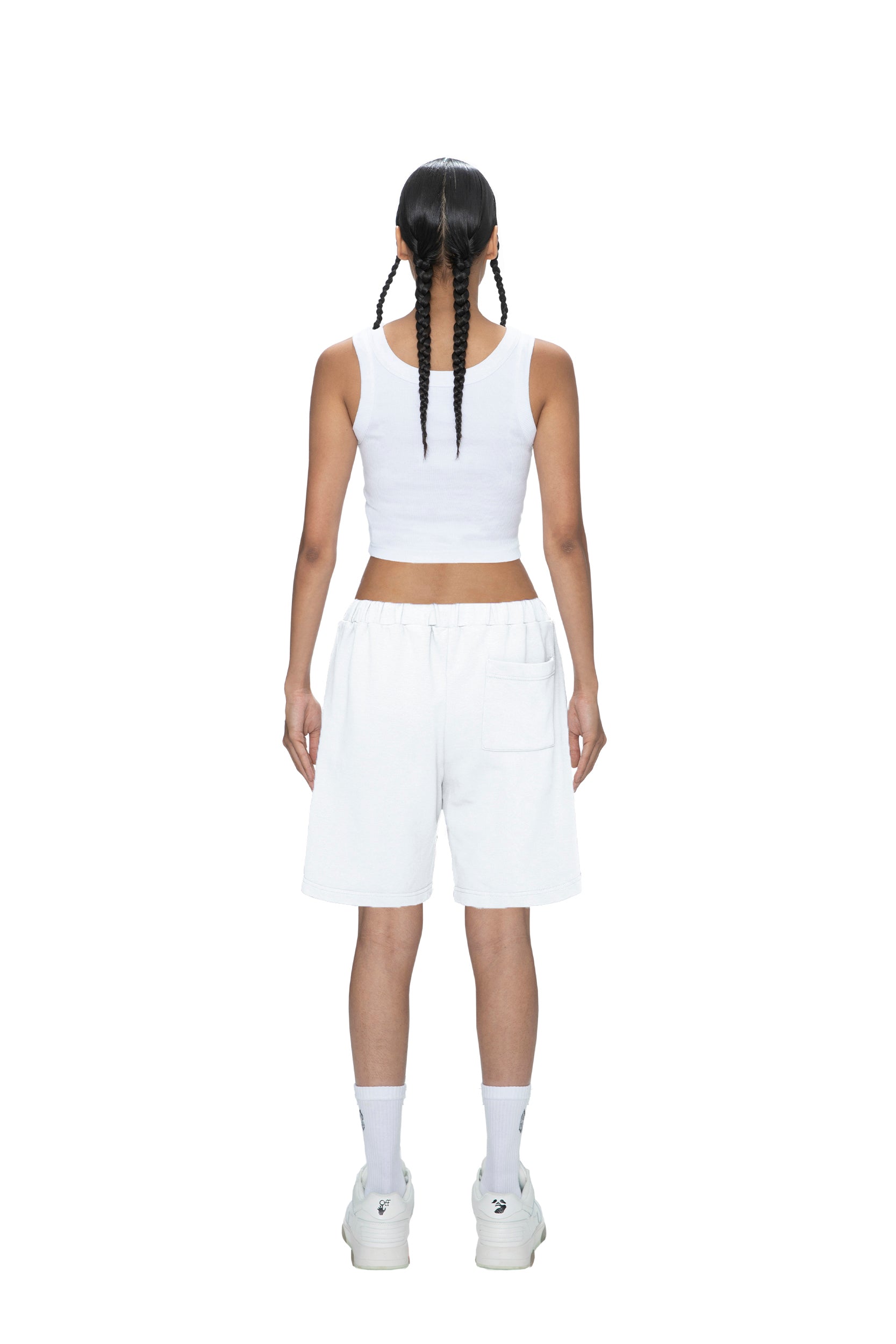 A person with two long braids stands facing away, wearing 6PM SWEATSHORTS WHITE, a white sleeveless crop top, white socks, and white sneakers against a plain white background.