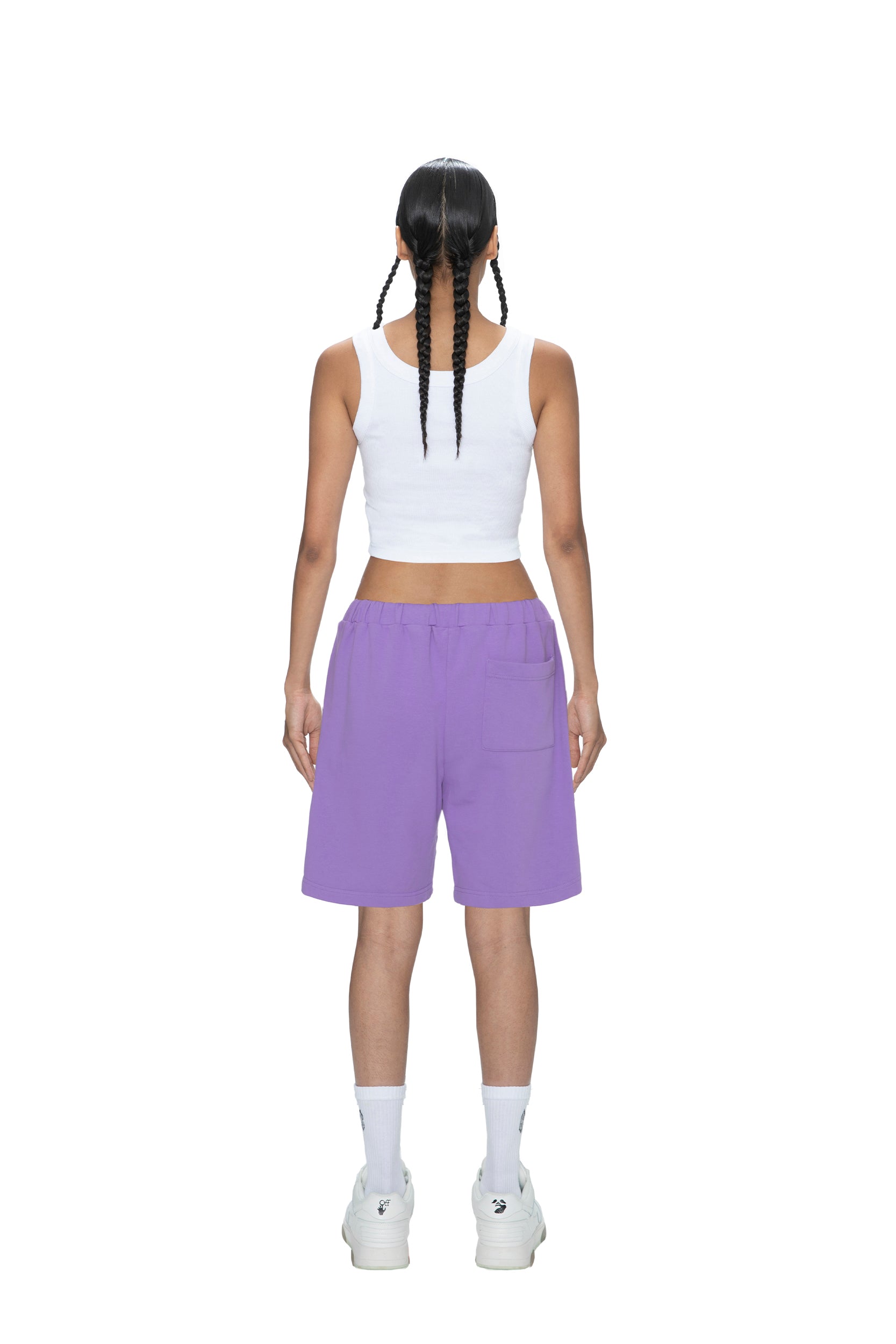 A person with two long braids wears a white sleeveless crop top, 6PM SWEATSHORTS LILAC, white socks, and white sneakers, standing facing away against a plain white background.