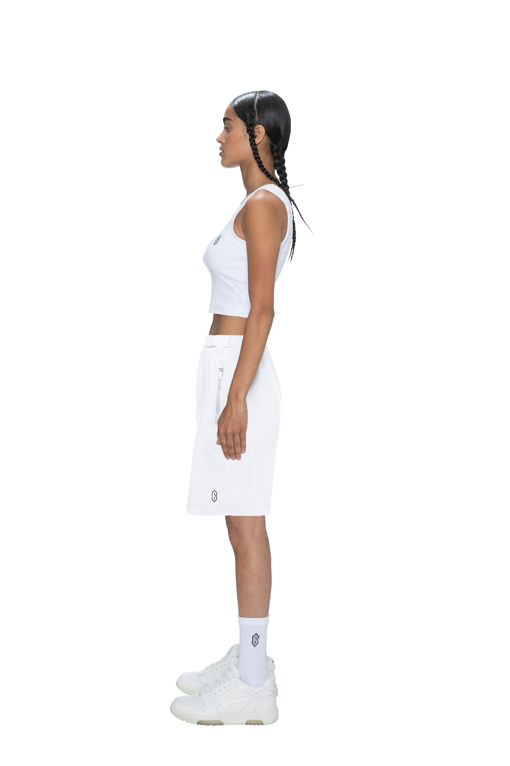 A woman with long braids stands in profile against a white background, wearing 6PM SWEATSHORTS WHITE along with a white crop top, white socks, and white sneakers.