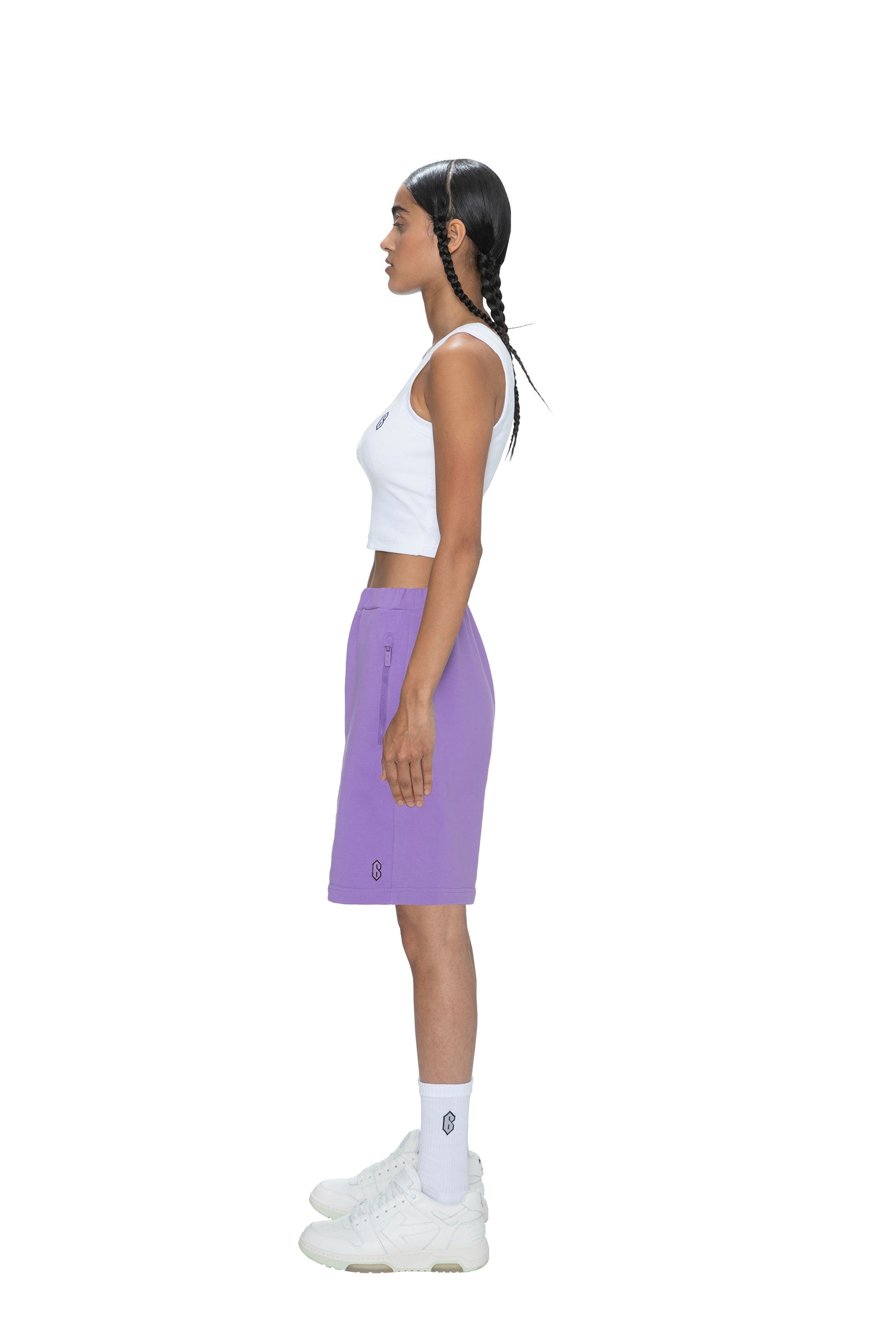 A woman with long braids stands in profile against a plain white background, wearing 6PM's SWEATSHORTS LILAC, a white crop top, white crew socks, and white sneakers.