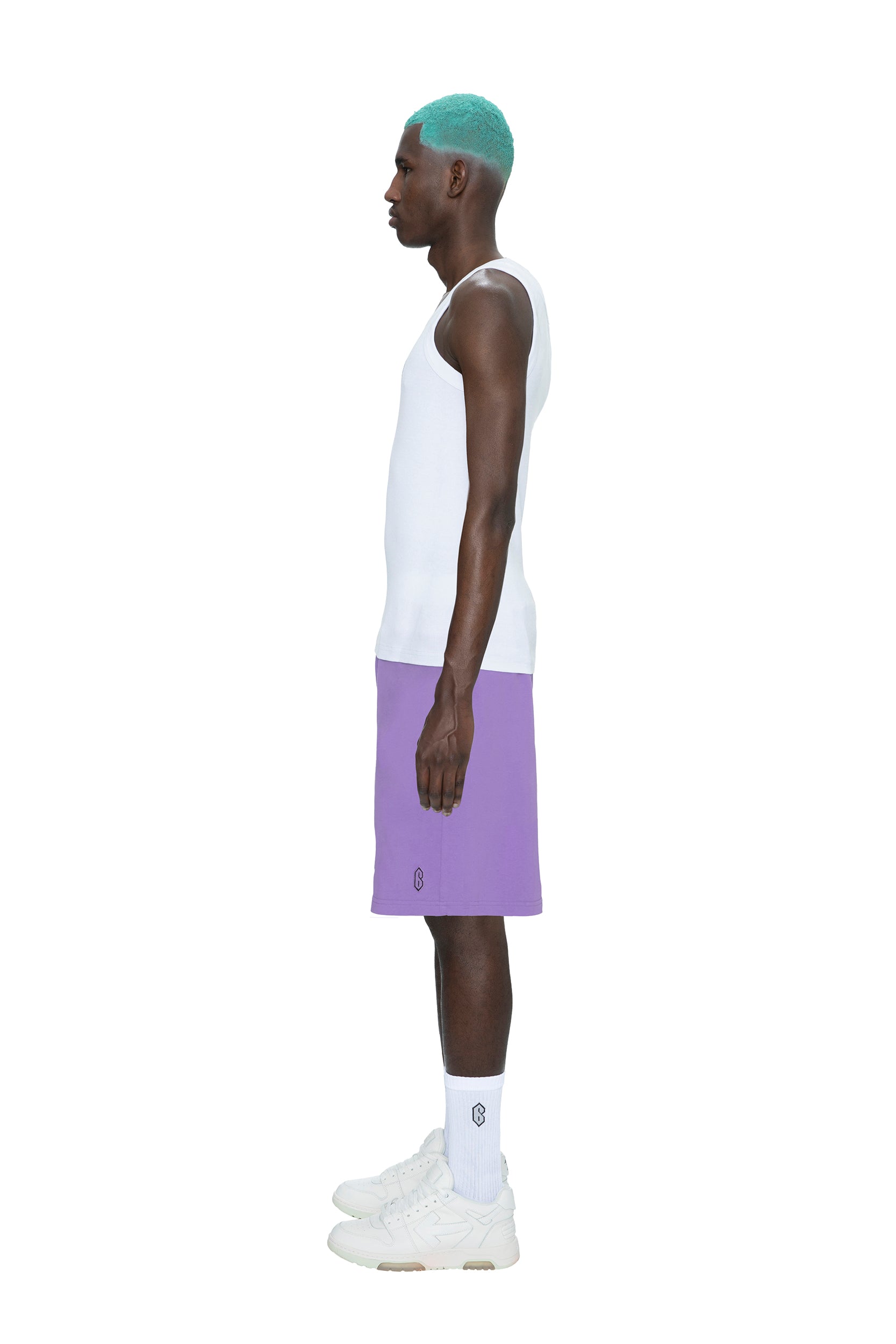 A man with teal hair stands in profile against a white background, wearing a white tank top, 6PM SWEATSHORTS LILAC, white crew socks, and white sneakers.