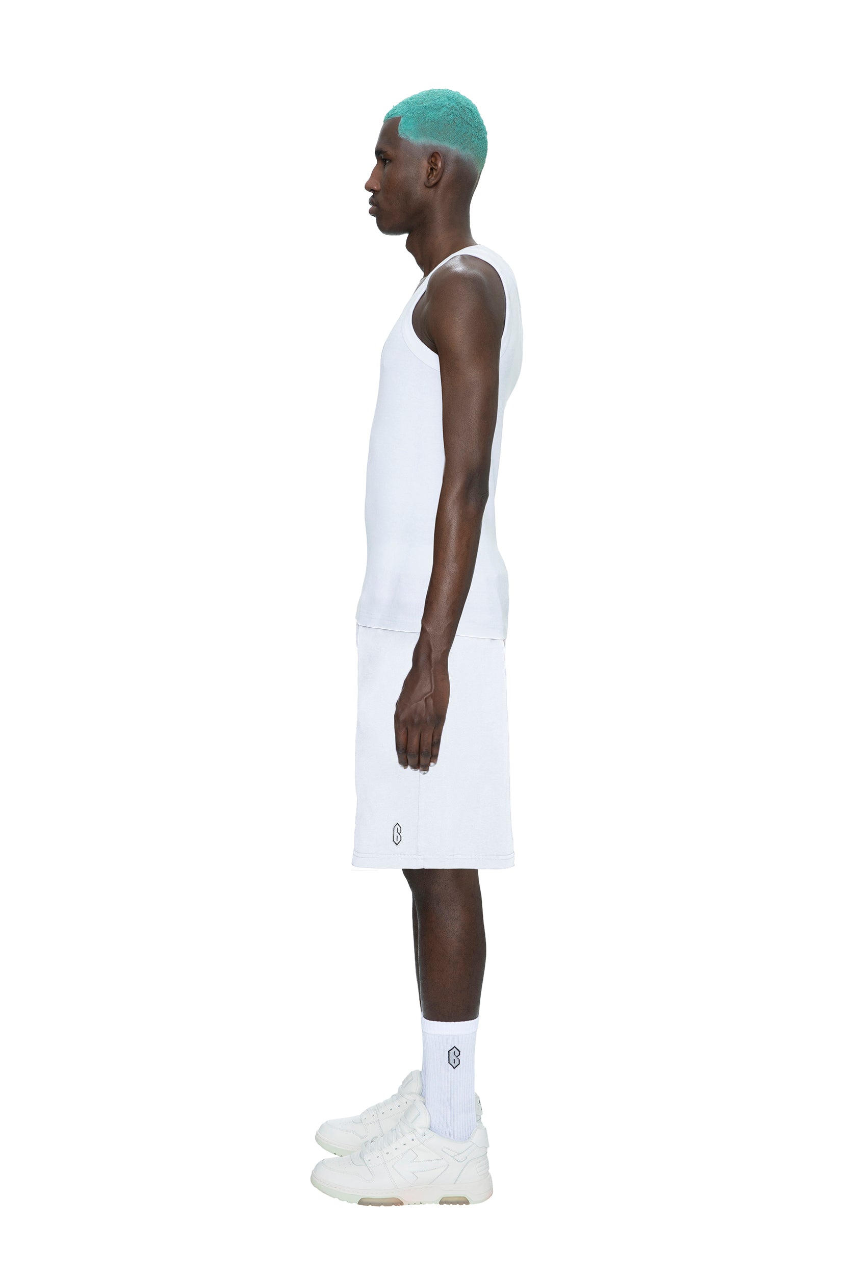 A man with turquoise hair stands in profile against a white background, wearing 6PM's SWEATSHORTS WHITE, a white tank top, white socks, and white sneakers.