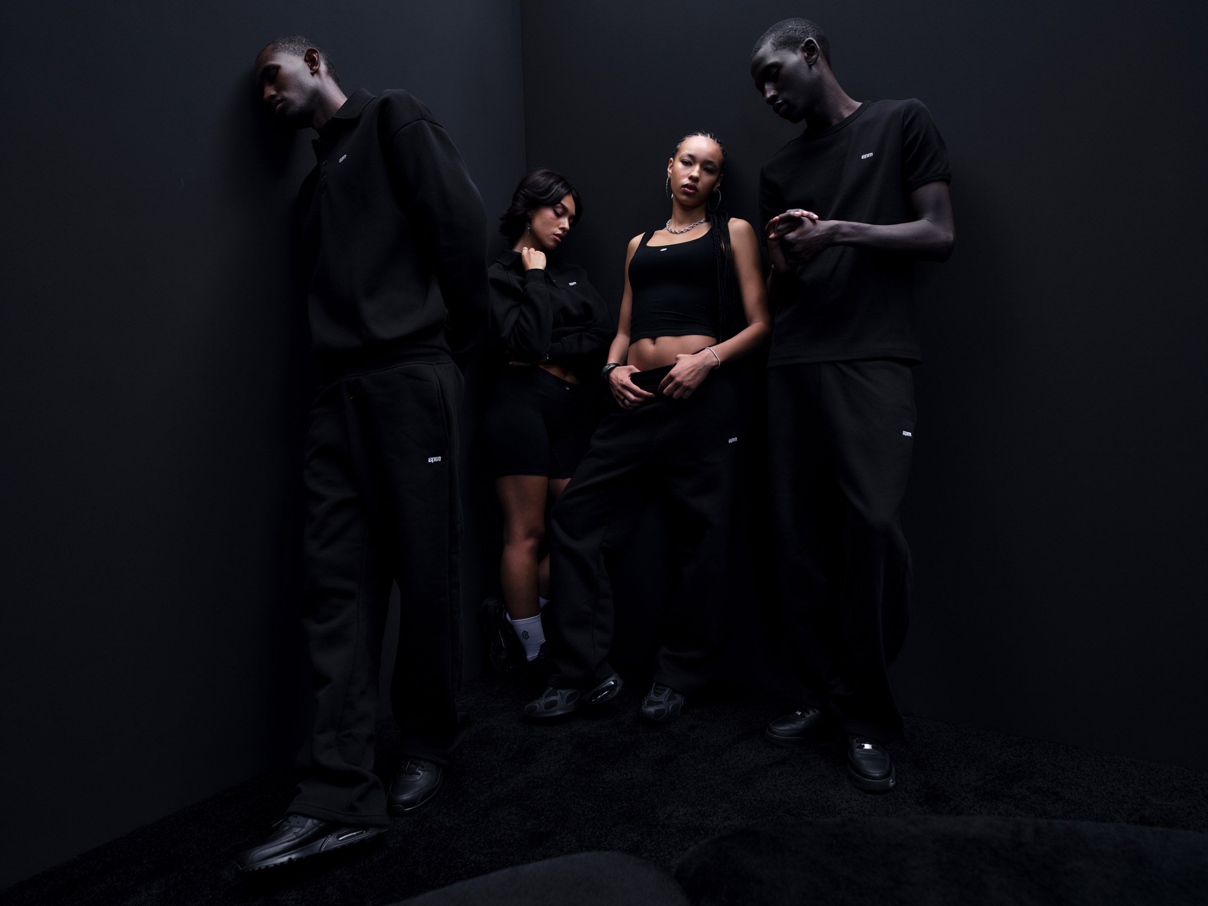 Four people dressed in all-black casual outfits stand together in a dimly lit corner, against dark walls and floor, creating a moody, minimalist vibe.
