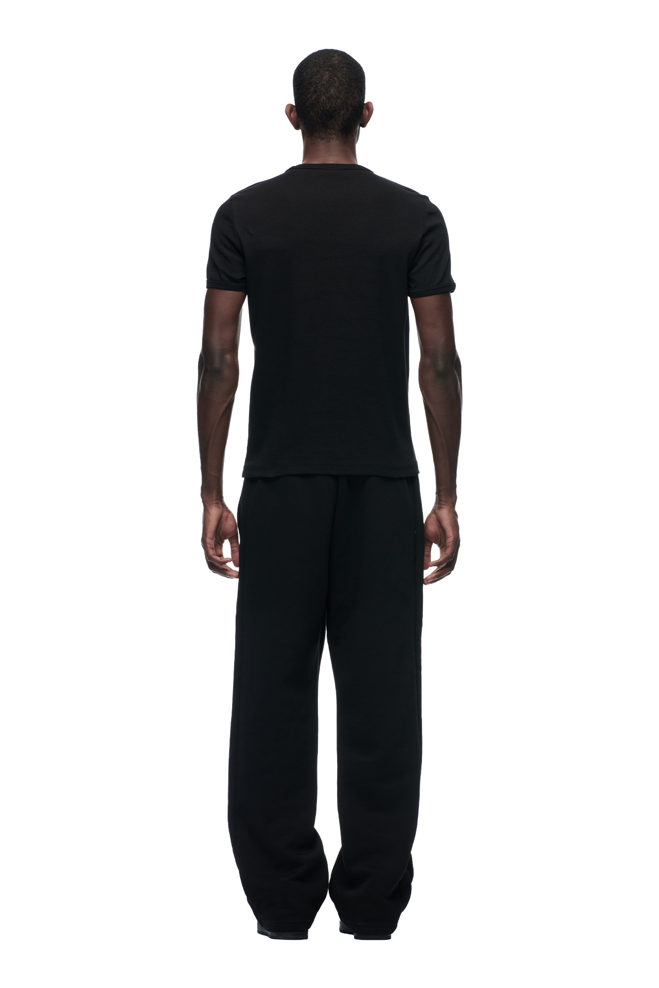 A man faces away from the camera wearing the 6PM Ribbed Slim Fit Tee Black and loose black pants, set against a plain white background.