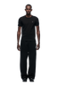 A man stands facing forward against a plain white background, wearing the 6PM RIBBED SLIM FIT TEE BLACK, loose black pants, and black shoes.