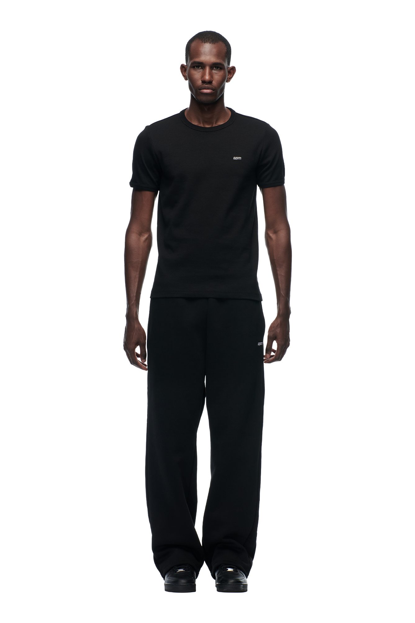 A man stands facing forward against a plain white background, wearing the 6PM RIBBED SLIM FIT TEE BLACK, loose black pants, and black shoes.
