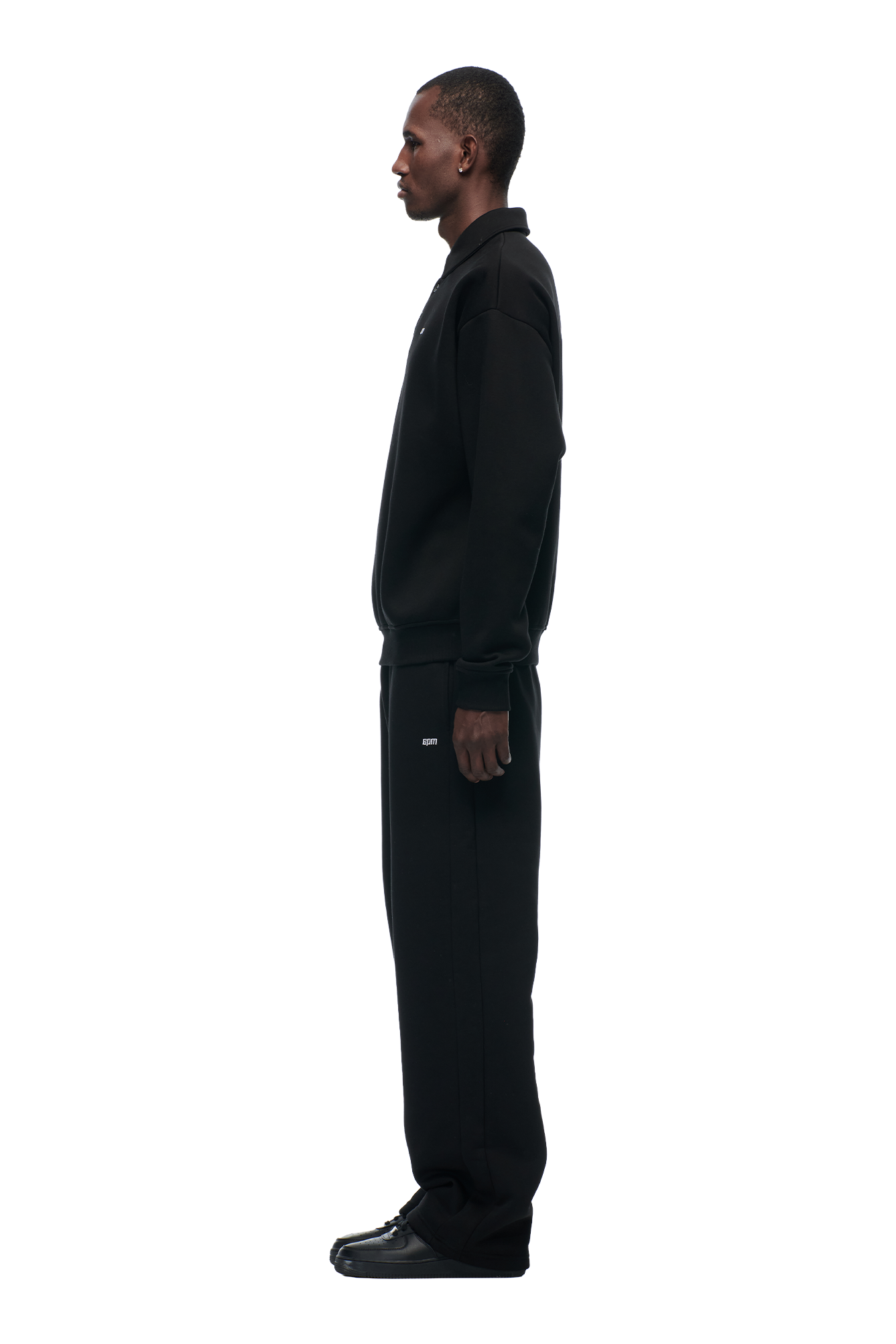A man stands in profile facing left, dressed in the POLO BLACK collared sweatshirt and matching sweatpants from 6PM with black sneakers, set against a plain white background.