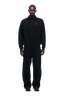 A man stands facing forward against a plain white background, wearing 6PM's POLO BLACK collared sweatshirt and matching loose black pants with black shoes.