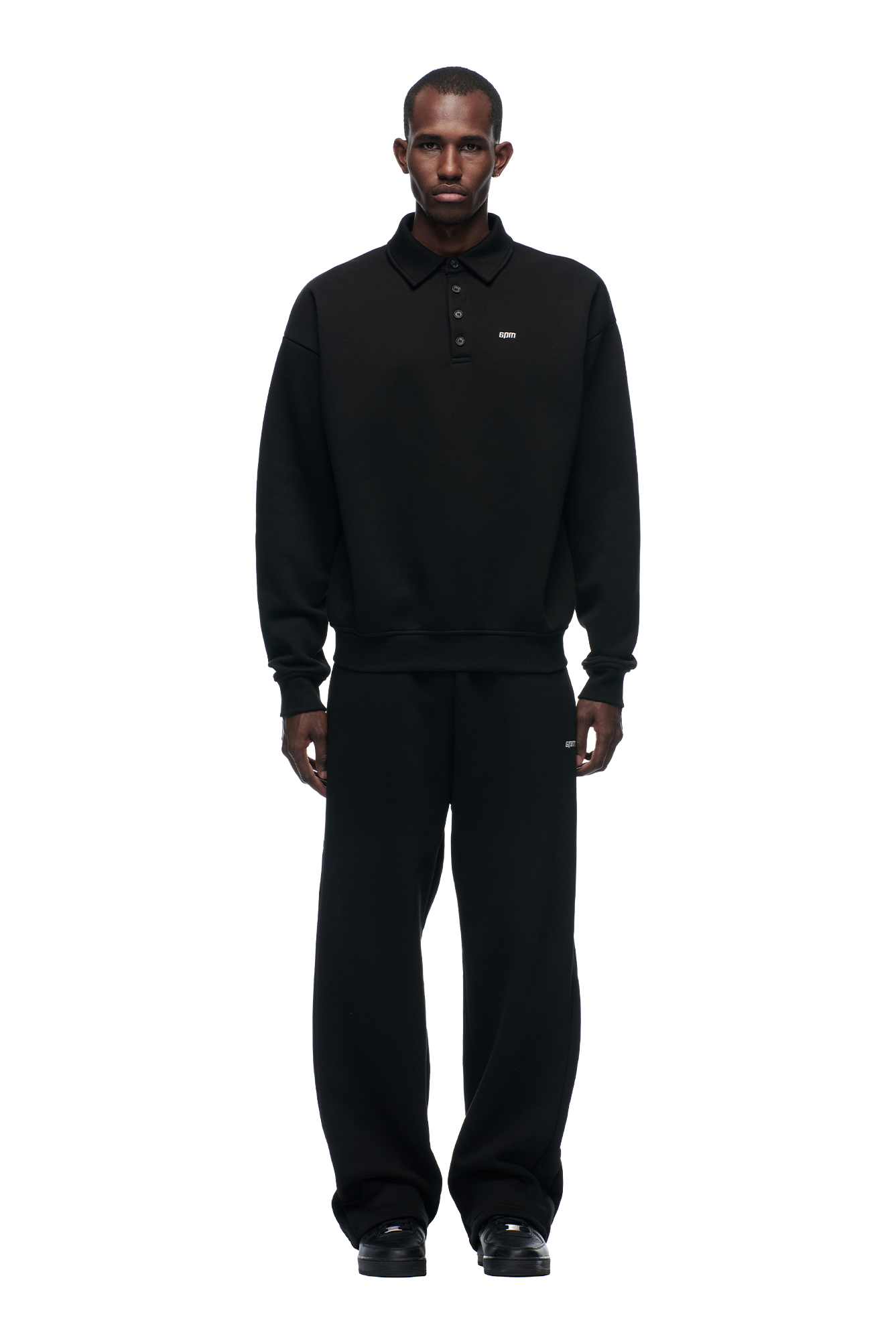 A man stands facing forward against a plain white background, wearing 6PM's POLO BLACK collared sweatshirt and matching loose black pants with black shoes.