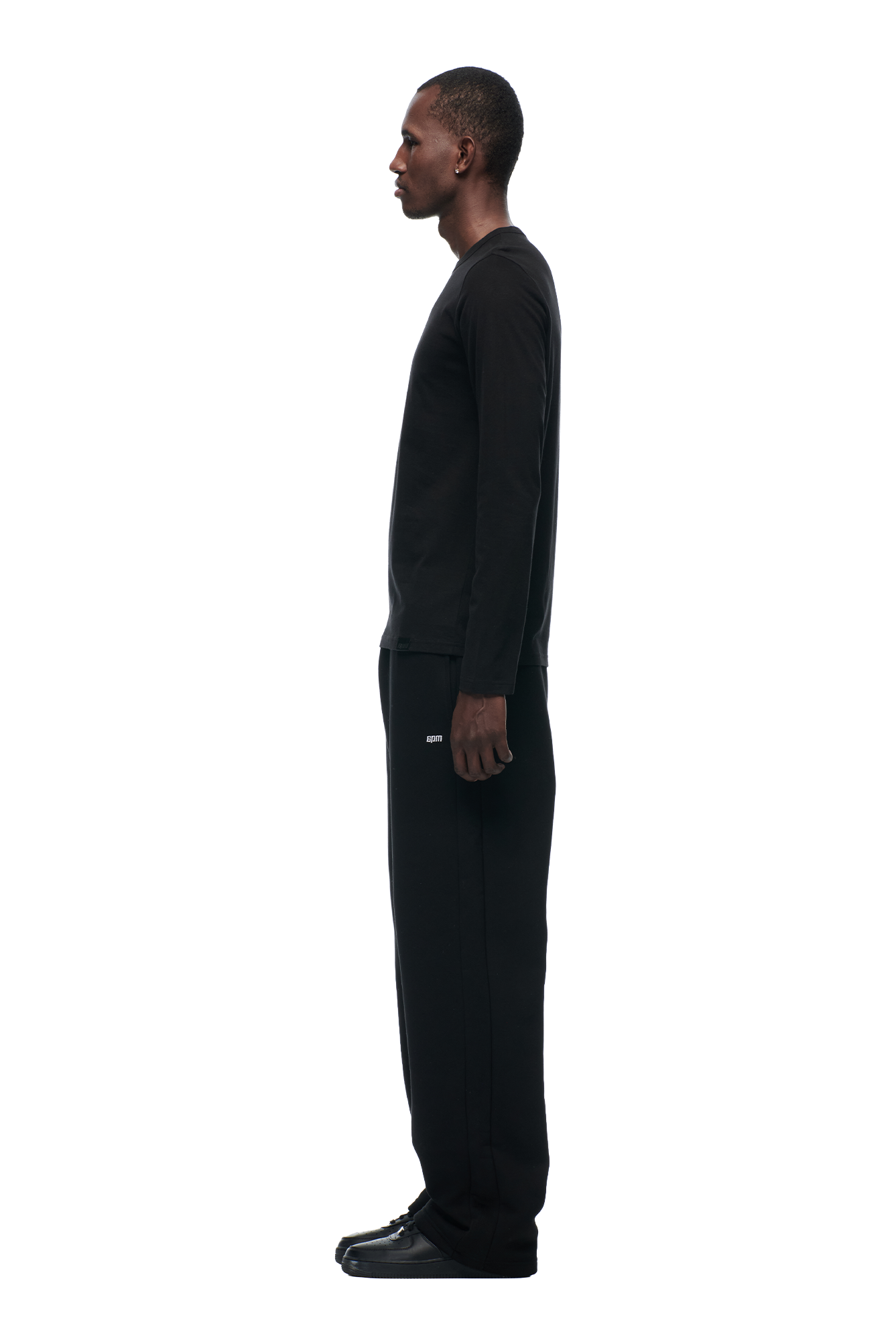 A man in profile facing left wears the 6PM CLEAN LONGSLEEVE BLACK with black pants and sneakers, posing against a plain white background.