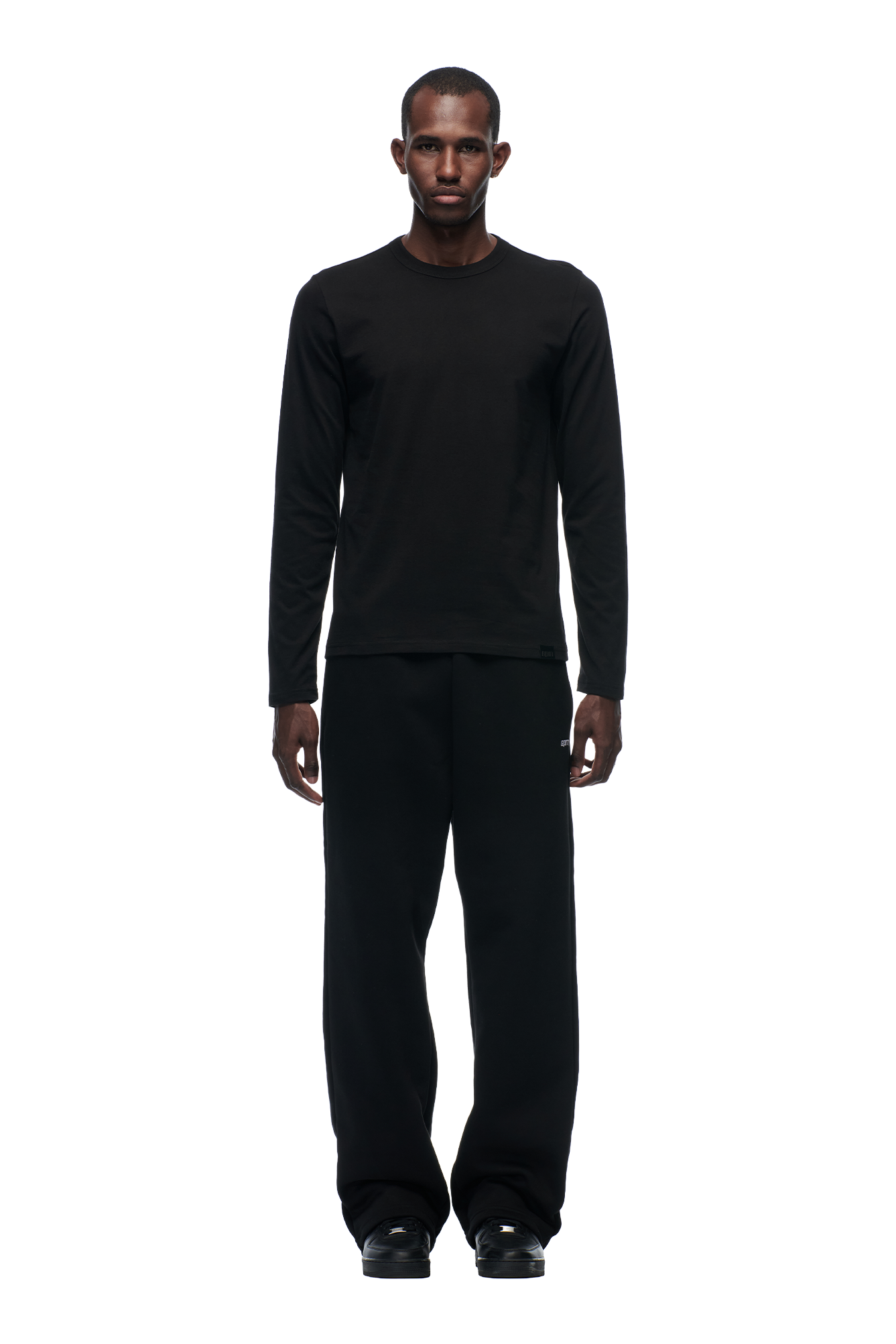 A man stands facing forward against a plain white background, wearing the 6PM CLEAN LONGSLEEVE BLACK shirt, along with black pants and shoes.