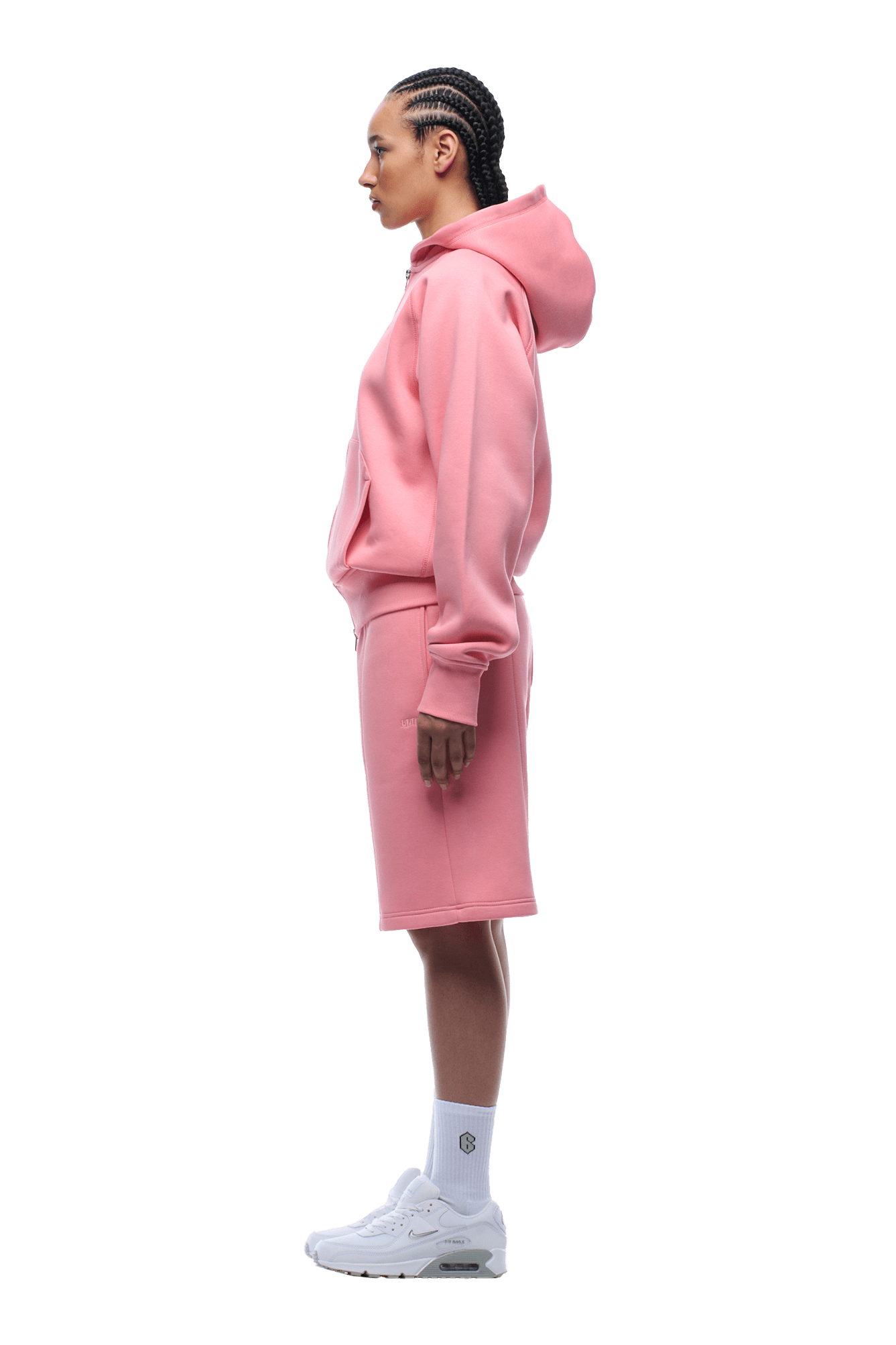 A person stands in profile wearing the 6PM COVERLOCK ZIP HOODIE FLAMINGO PINK, matching knee-length shorts, white crew socks, and white sneakers. They have braided hair and a neutral expression against a transparent background.