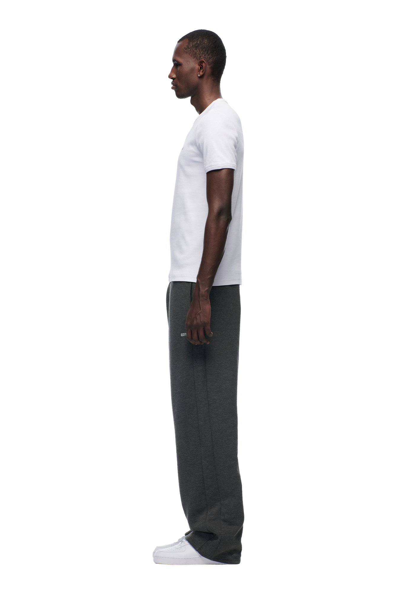A man stands in profile against a plain white background, wearing the 6PM RIBBED SLIM FIT TEE WHITE, paired with dark gray sweatpants and white sneakers.