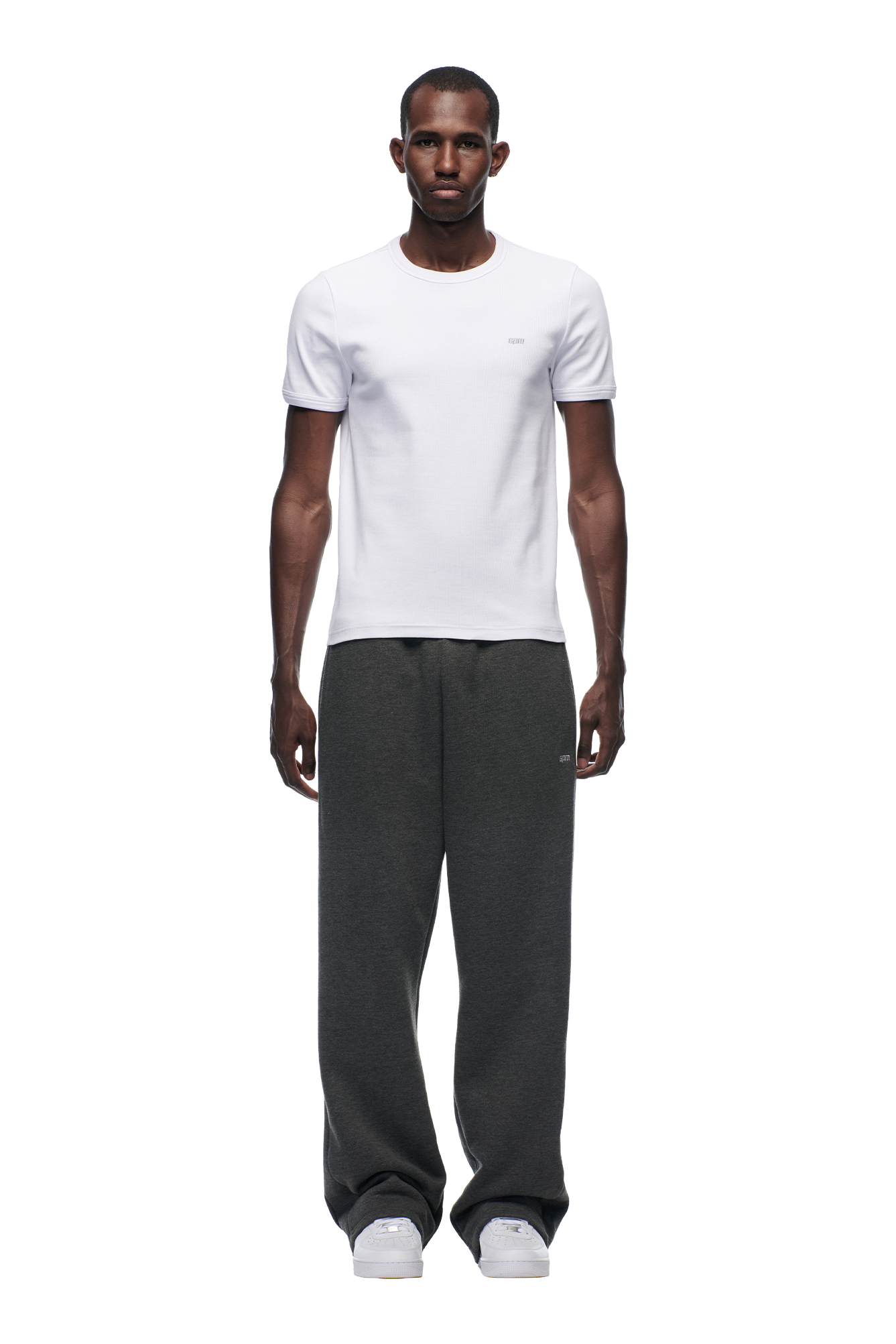 A man faces forward against a plain white background, wearing the 6PM RIBBED SLIM FIT TEE in white, loose dark gray sweatpants, and white sneakers.