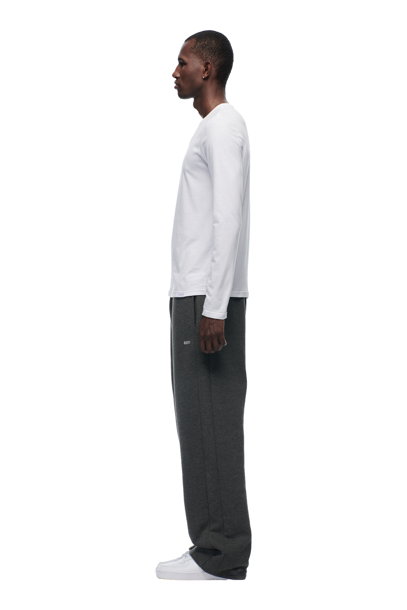 A man stands in profile facing left, wearing the 6PM CLEAN LONGSLEEVE WHITE shirt, dark gray sweatpants, and white sneakers against a plain white background.