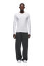 A man stands facing forward, wearing the 6PM CLEAN LONGSLEEVE WHITE shirt, loose dark gray sweatpants, and white sneakers against a white background.