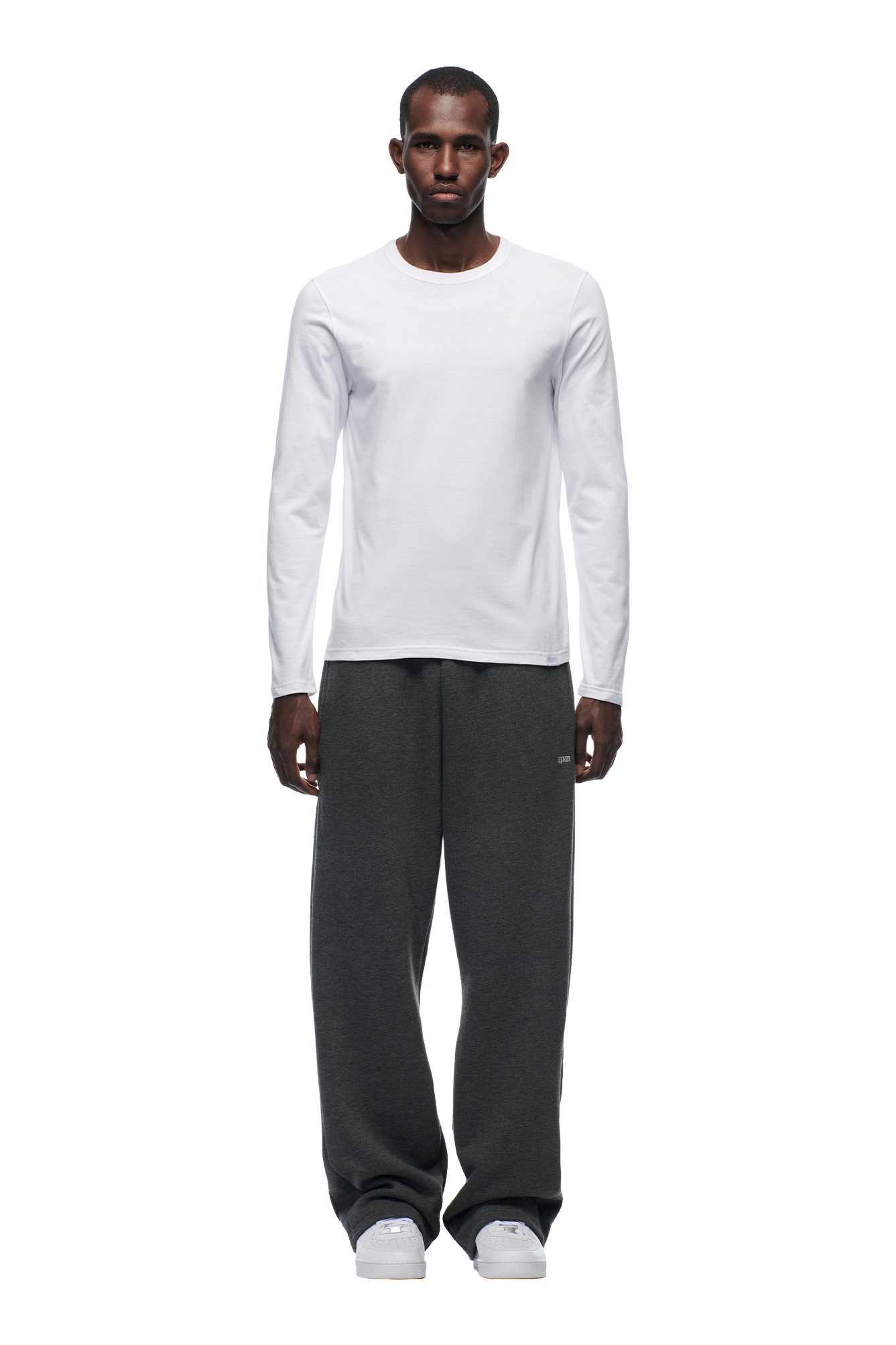 A man stands facing forward, wearing the 6PM CLEAN LONGSLEEVE WHITE shirt, loose dark gray sweatpants, and white sneakers against a white background.