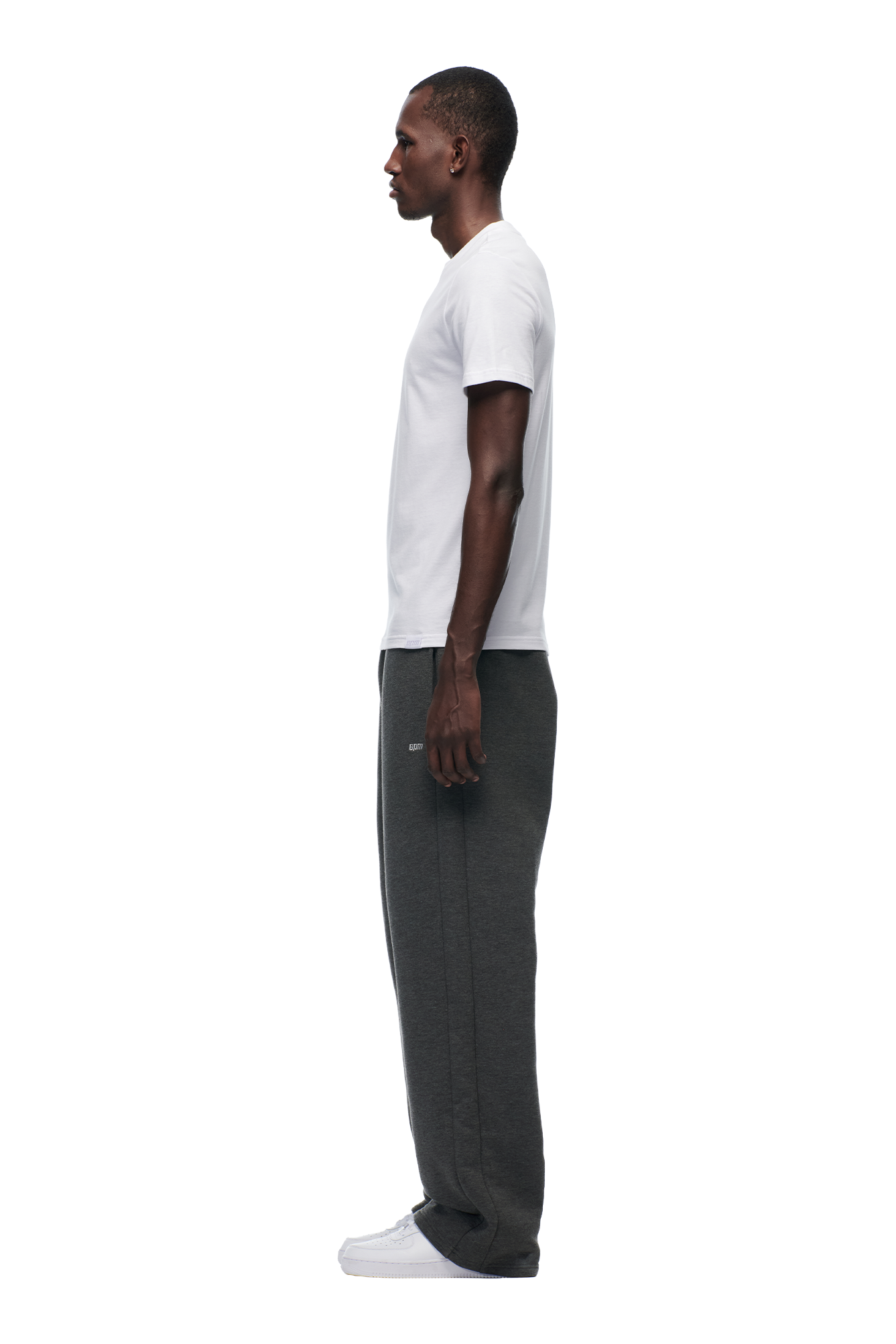 A man in profile faces left, wearing the 6PM SLIM FIT T-SHIRT WHITE in premium cotton, paired with dark gray sweatpants and white sneakers against a plain light background.