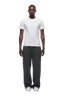 A man stands facing forward wearing the 6PM SLIM FIT T-SHIRT WHITE, paired with loose-fitting dark gray sweatpants and white sneakers, set against a plain white background.