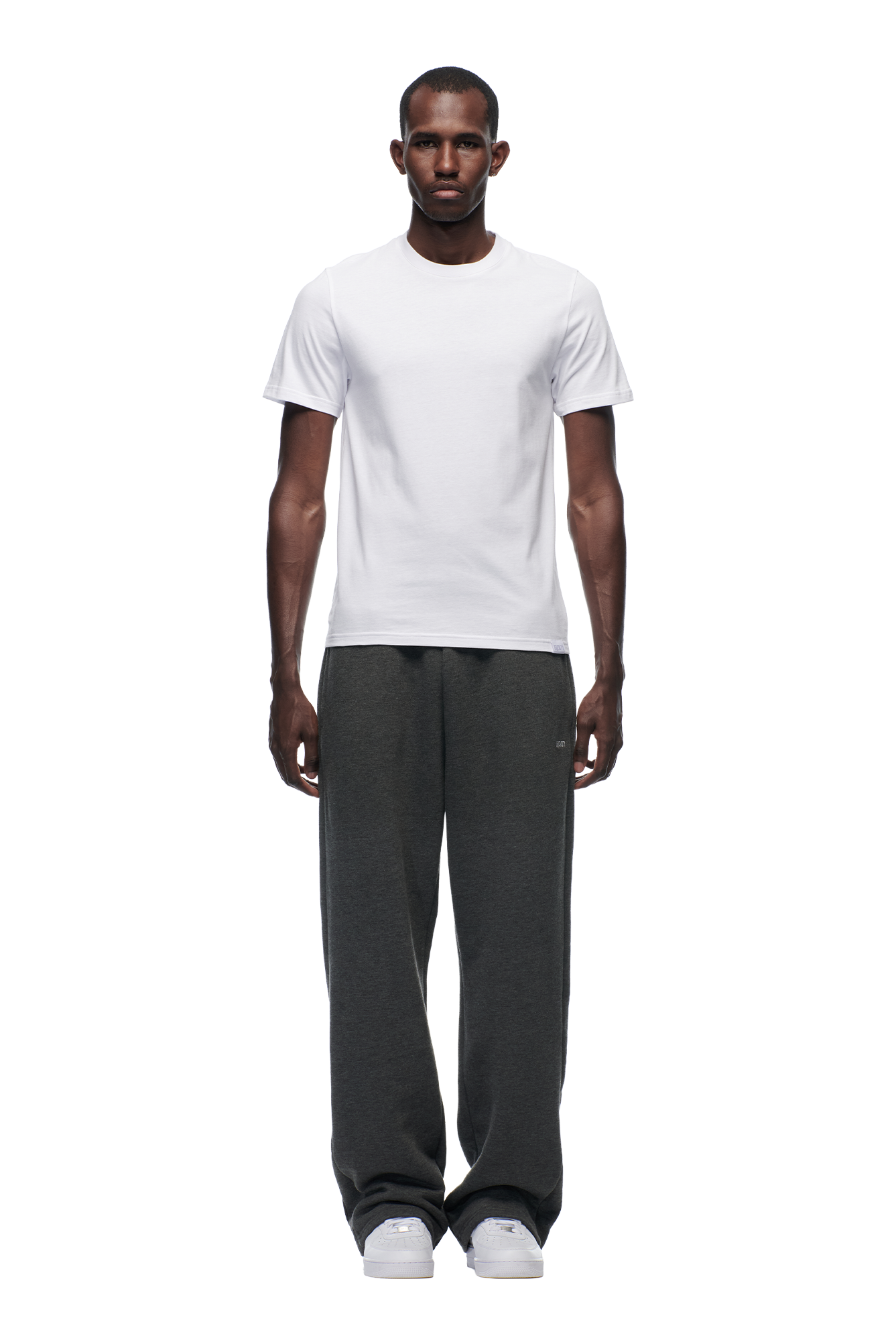 A man stands facing forward wearing the 6PM SLIM FIT T-SHIRT WHITE, paired with loose-fitting dark gray sweatpants and white sneakers, set against a plain white background.