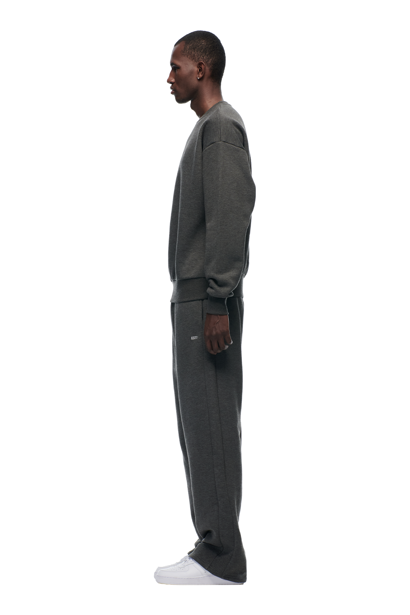 A person stands in profile facing left, wearing the 6PM CREWNECK DARK GREY MELANGE sweatshirt and matching sweatpants with white sneakers, against a plain light gray background.
