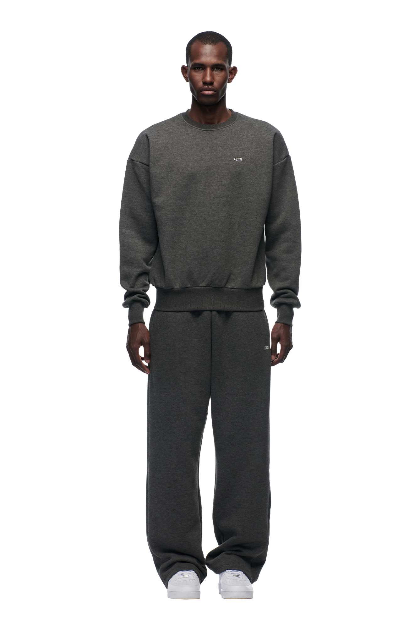 A man stands facing forward wearing the 6PM CREWNECK DARK GREY MELANGE sweatshirt with ribbed trims, matching sweatpants, and white sneakers against a plain white background, his hands resting by his sides.