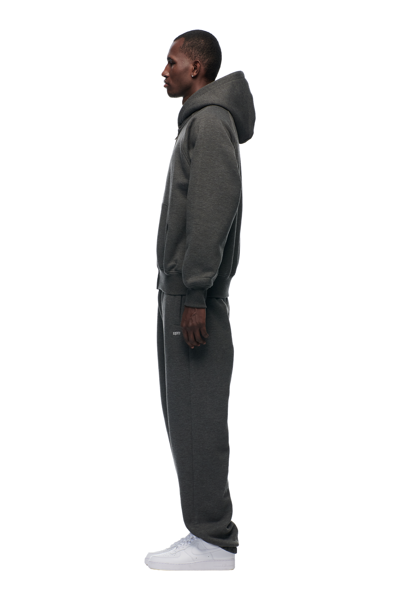 A person stands in profile against a plain background, wearing the 6PM ZIP HOODIE DARK GREY MELANGE with matching sweatpants and white sneakers.