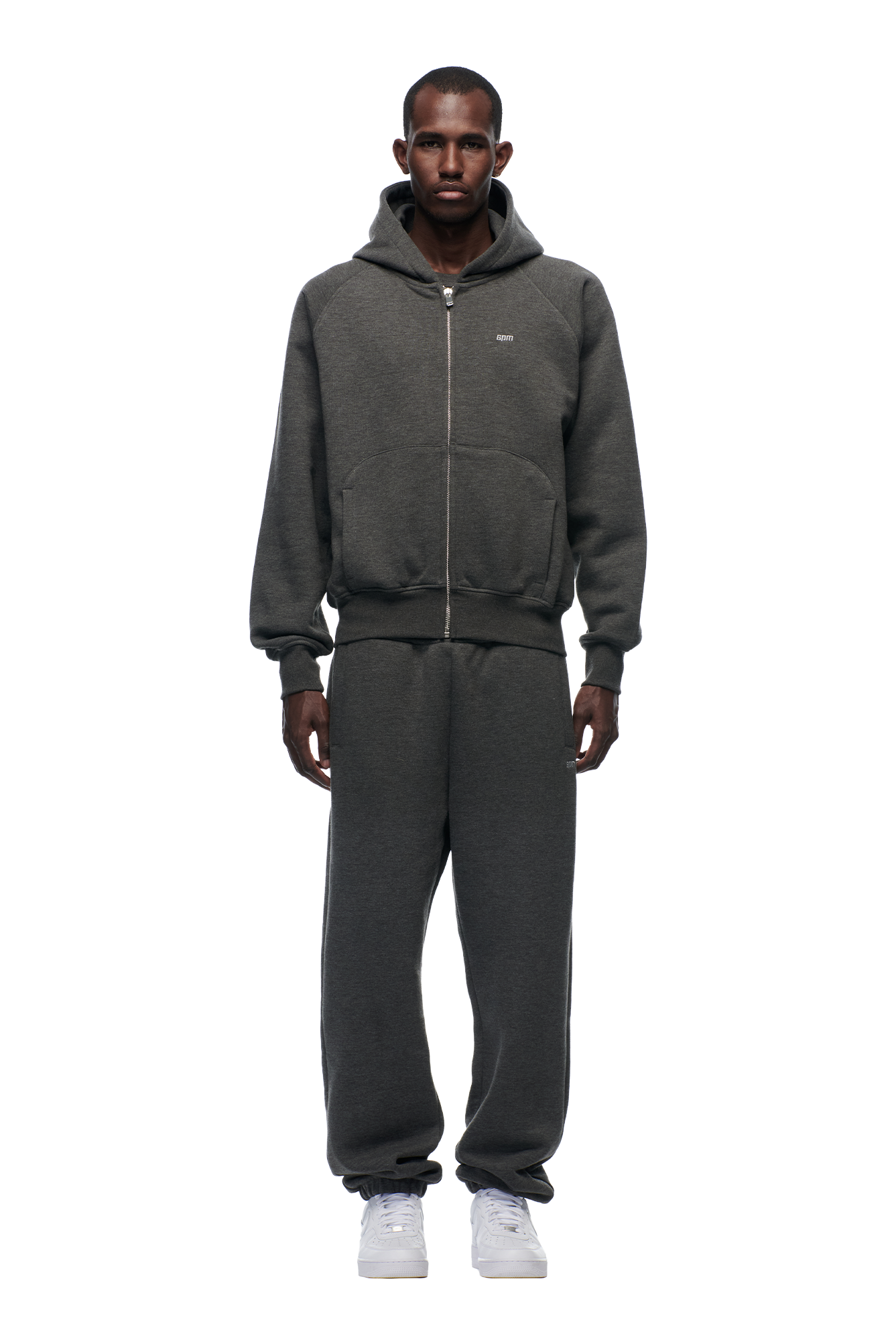 A man faces forward against a plain white background, wearing the 6PM ZIP HOODIE DARK GREY MELANGE with matching sweatpants and white sneakers.