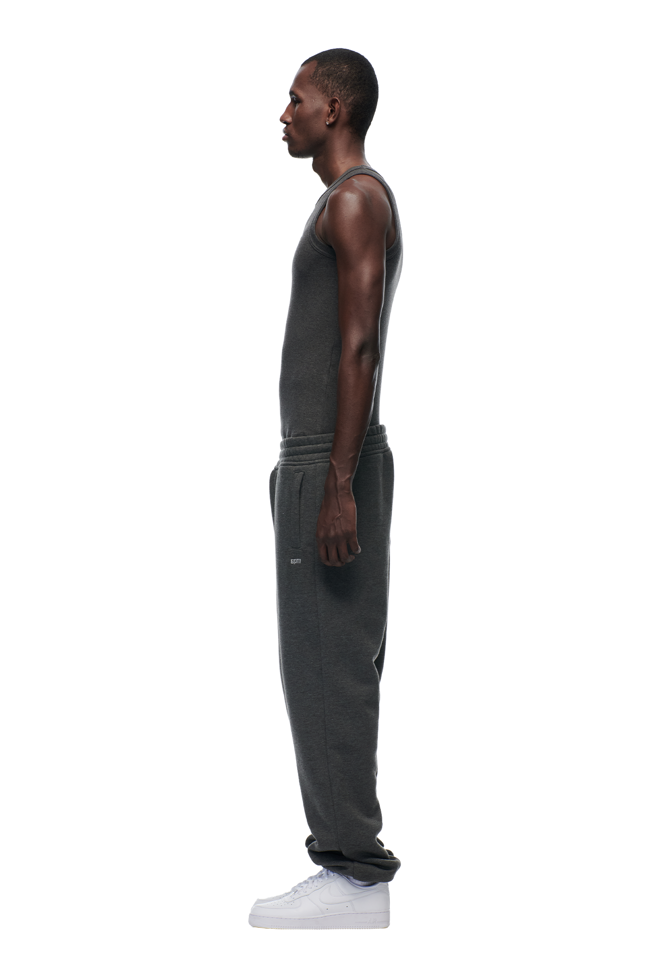 A man in profile faces left, wearing 6PM's CUFF JOGGER DARK GREY MELANGE sweatpants and a dark gray tank top with white sneakers, set against a plain white background.