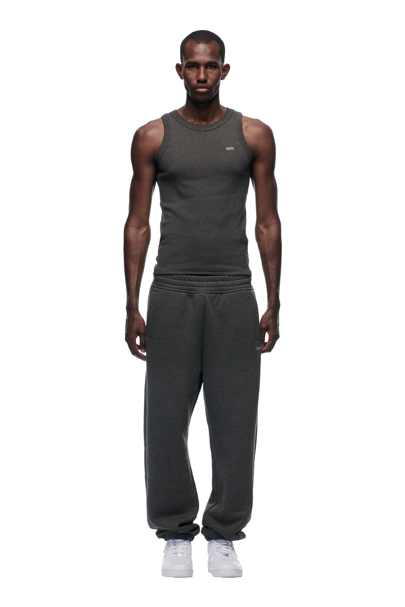 A man faces forward against a plain white background, wearing the 6PM CUFF JOGGER DARK GREY MELANGE sweatpants with a matching dark gray sleeveless tank top and white sneakers.