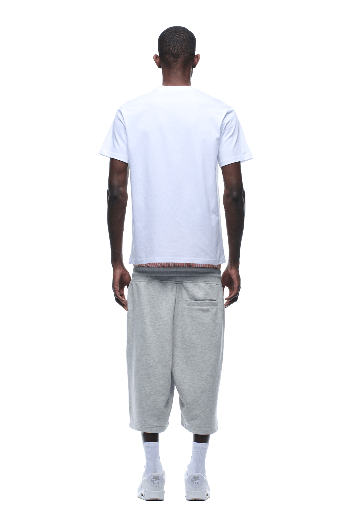 A man faces away from the camera wearing the 6PM CLEAN EDGE WAVE TEE in white, paired with light gray knee-length shorts, white socks, and sneakers, arms relaxed by his sides against a plain background.